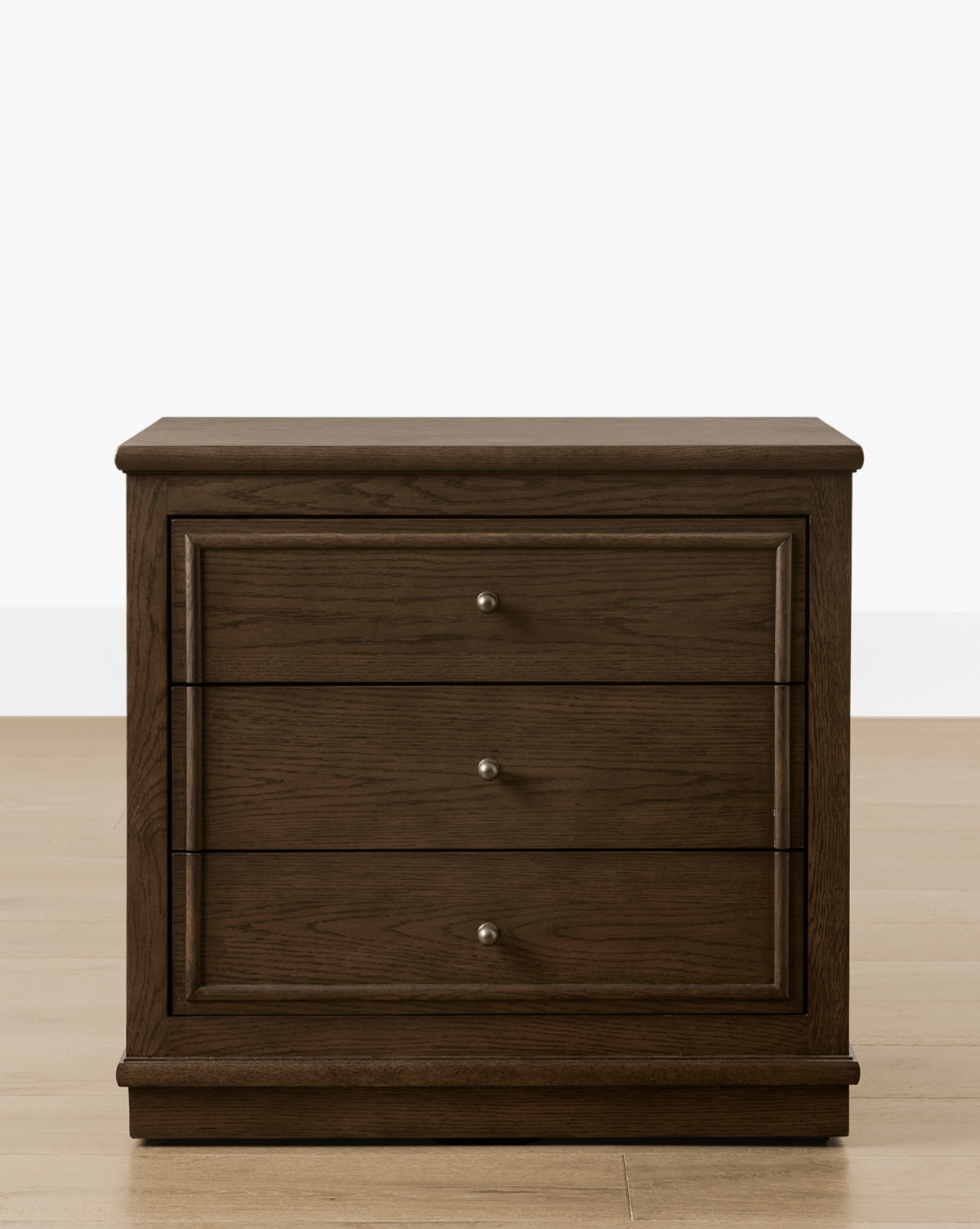 The Yorkshire Nightstand showcases a rich English oak finish with three drawers featuring antique brass pulls, elegantly placed on a light wood floor against a plain white background, by McGee & Co.