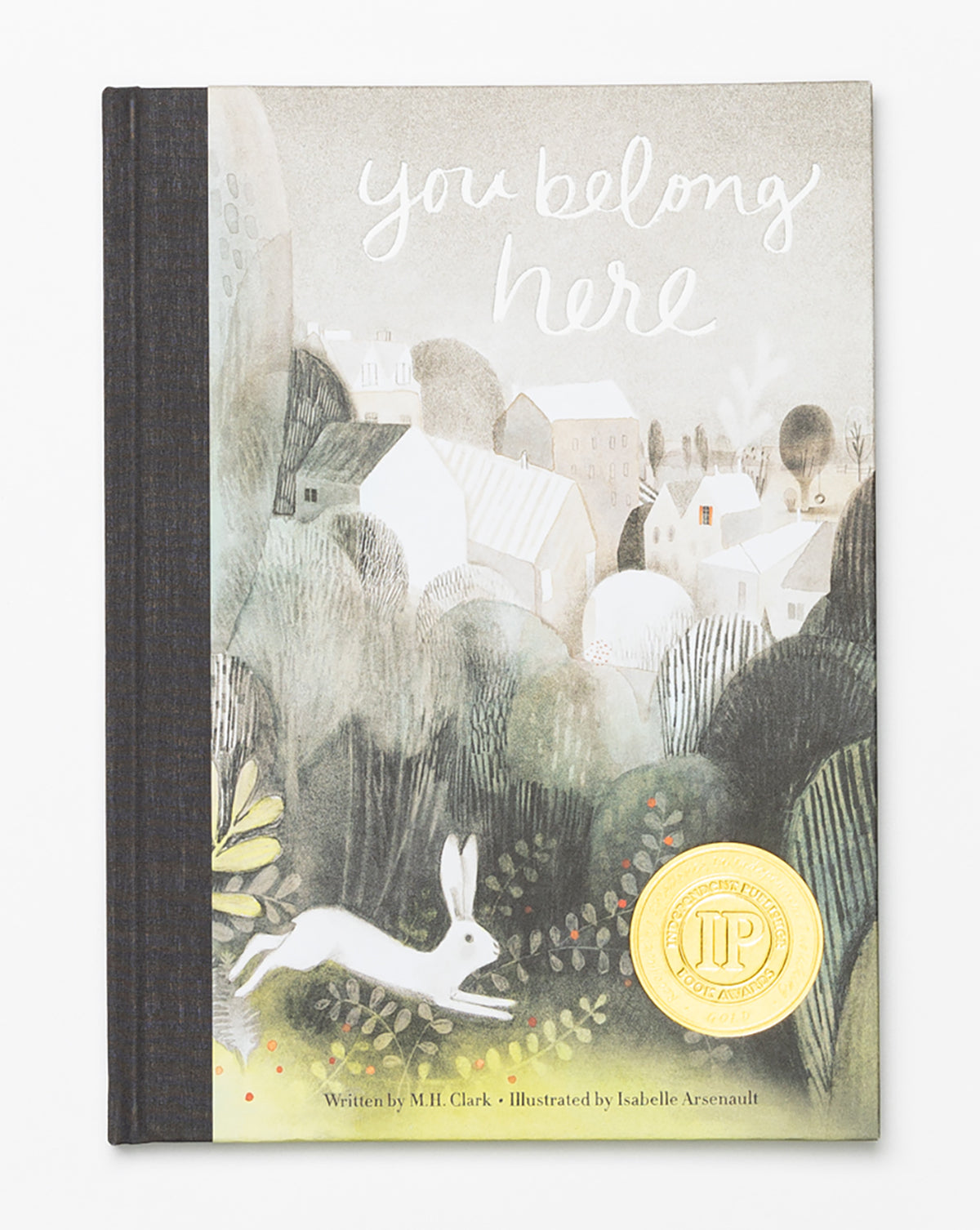 The childrens book You Belong Here features a whimsical white rabbit, green foliage, a village of light-colored houses, an award medal on the cover, and shares a heartfelt message of belonging.