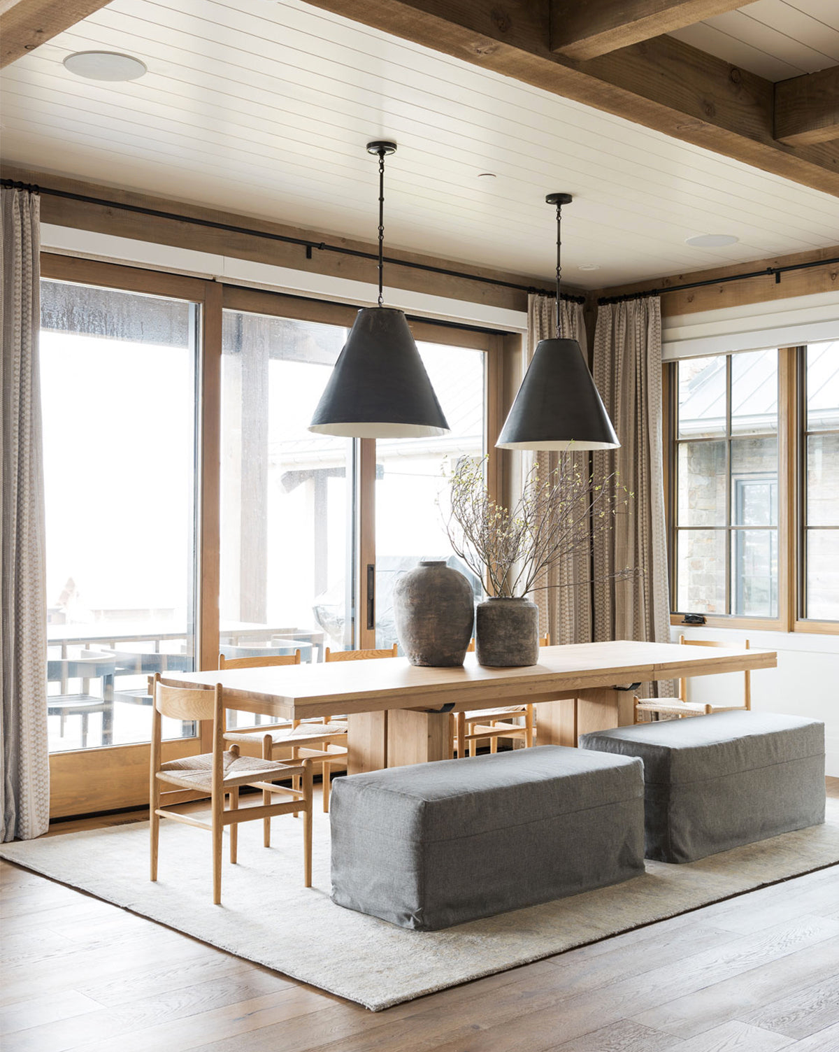 A modern dining room features the Antoni Extension Dining Table, six wooden chairs, two gray cushioned benches, black pendant lights, and vases with branches. Large windows and light curtains fill the space with natural light.