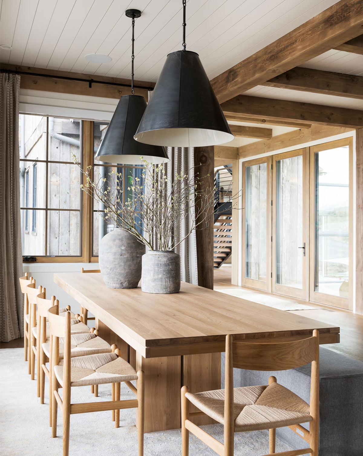 A modern rustic dining room features the Antoni Extension Dining Table with eight wooden chairs, two large ceramic vases filled with branches, oversized black pendant lights, and large windows that fill the space with natural light.