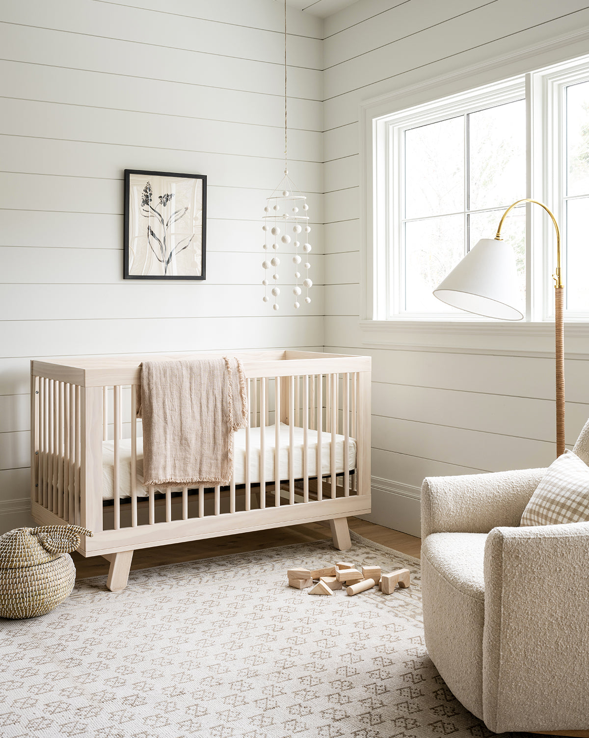 Bright, cozy nursery with a light wood crib, cream blanket, soft armchair, and the Anya Neutral Hand-Knotted Wool Rug. Wooden blocks, a basket, mobile, and botanical wall art add modern touches as sunlight streams through a large window, by McGee & Co.