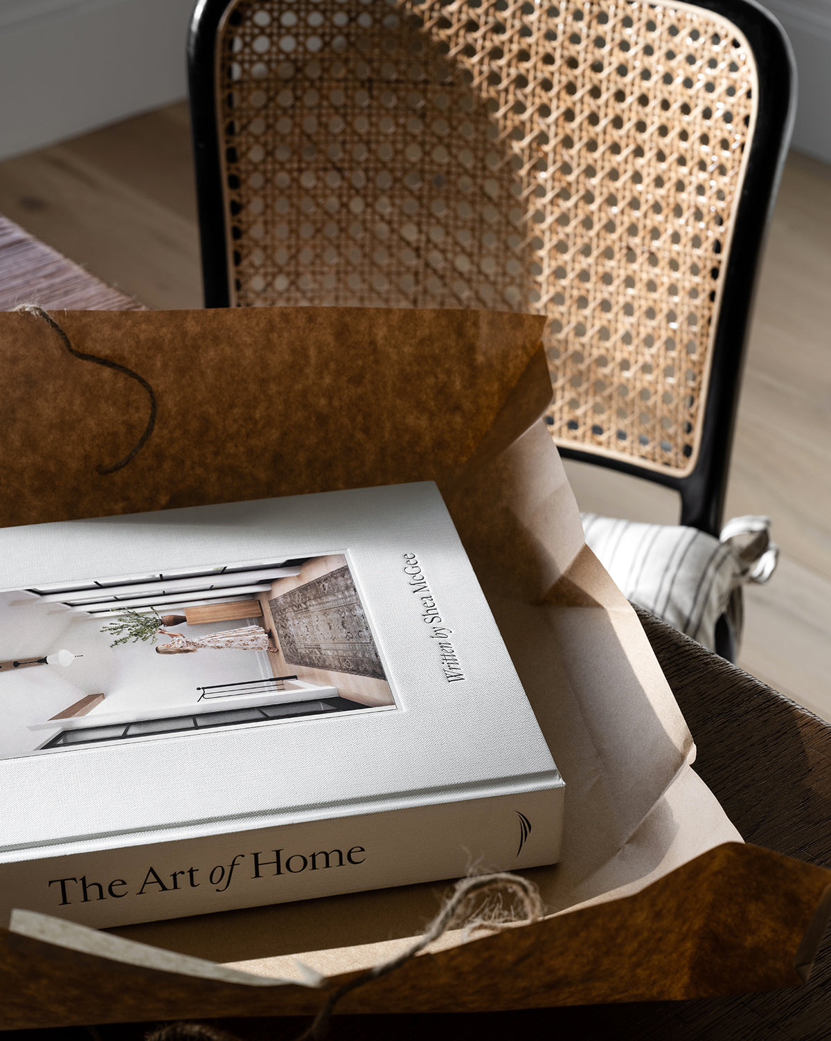 The Art of Home book, partially unwrapped on brown paper, offers interior design inspiration as it rests before a sunlit woven cane chair with a black wooden frame.