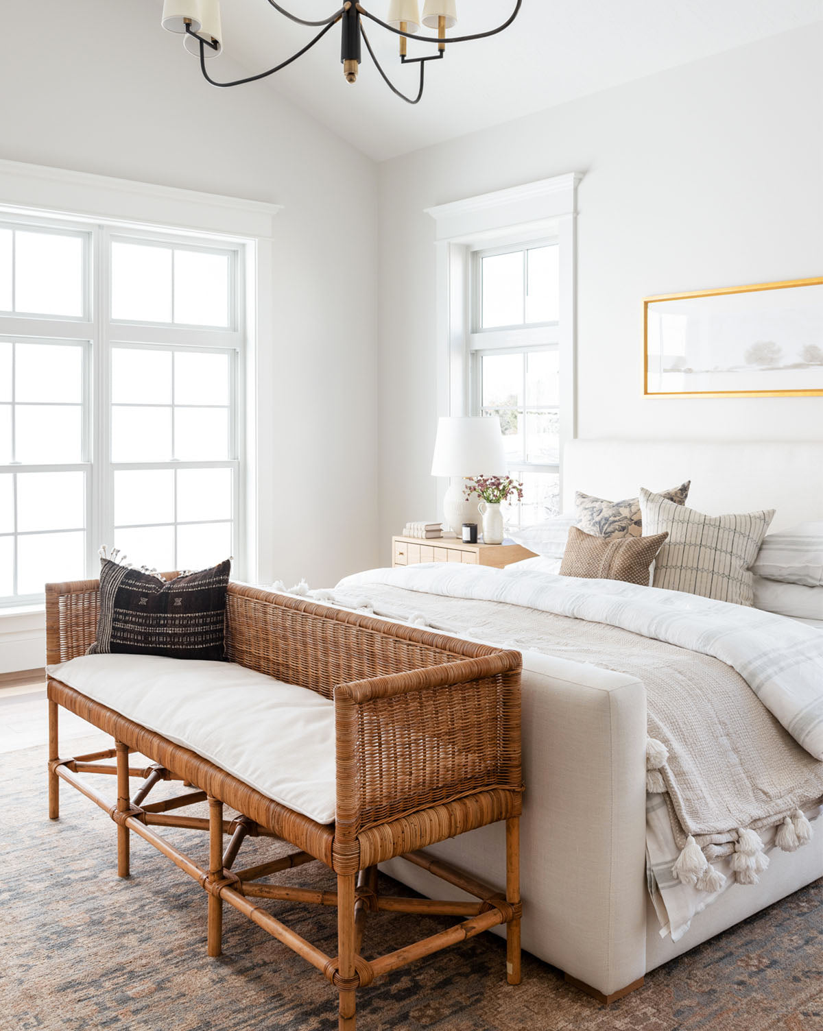 A bright, airy bedroom with large windows, a white bed adorned with performance fabric pillows and throws, the Odelia Bench at the foot, a bedside table with a white lamp and flowers, and framed art above the bed.