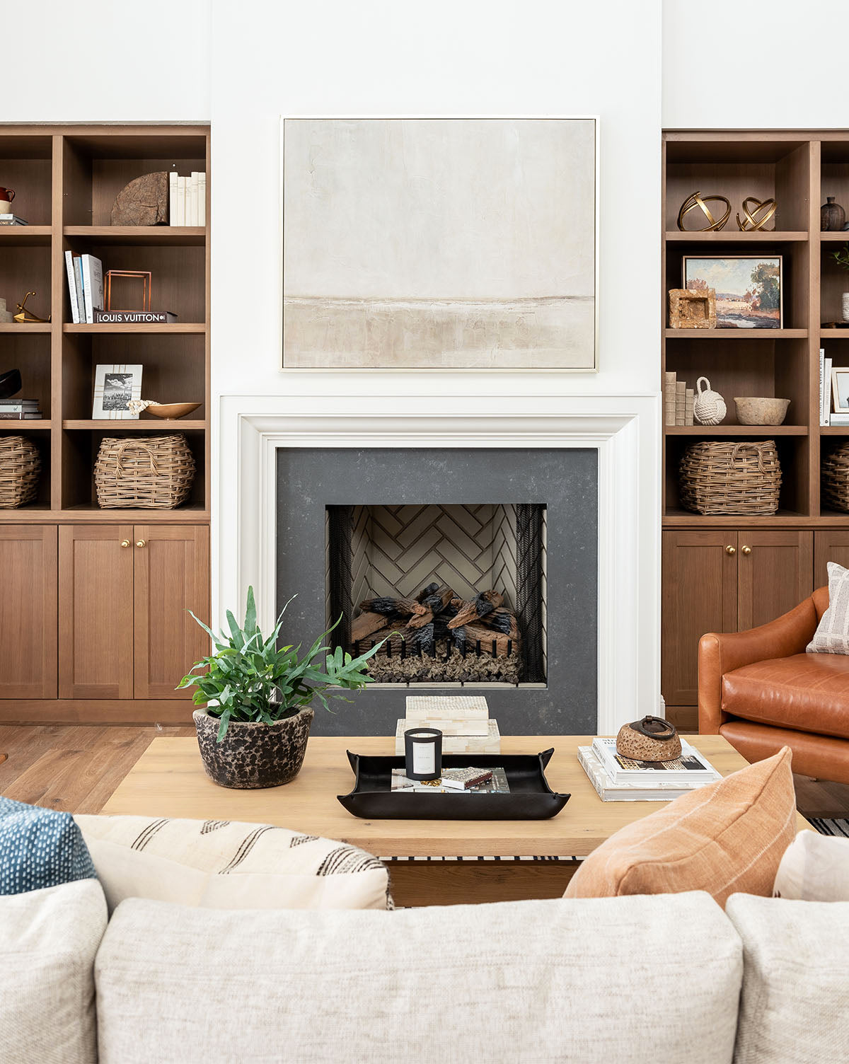 Create a breezy Gulf Shores vibe with a white sofa, tan leather chair, modern fireplace, and decor in gray-green hues. Wooden shelves show books, wicker baskets, a potted plant, and neutral abstract art above the mantel.