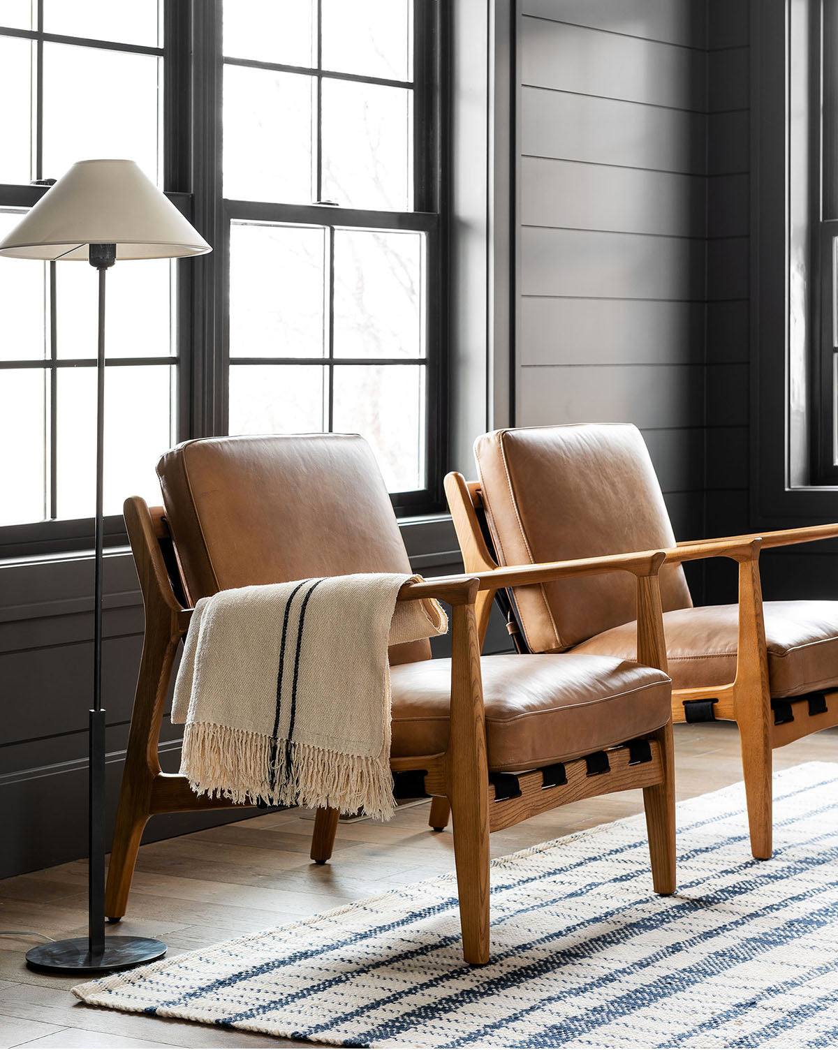 Two Lanston Lounge Chairs with ash wood frames and tan leather cushions sit by large windows. One is draped with a striped throw blanket, while a tall floor lamp and blue-and-white striped rug complete the cozy, modern setting from McGee & Co,