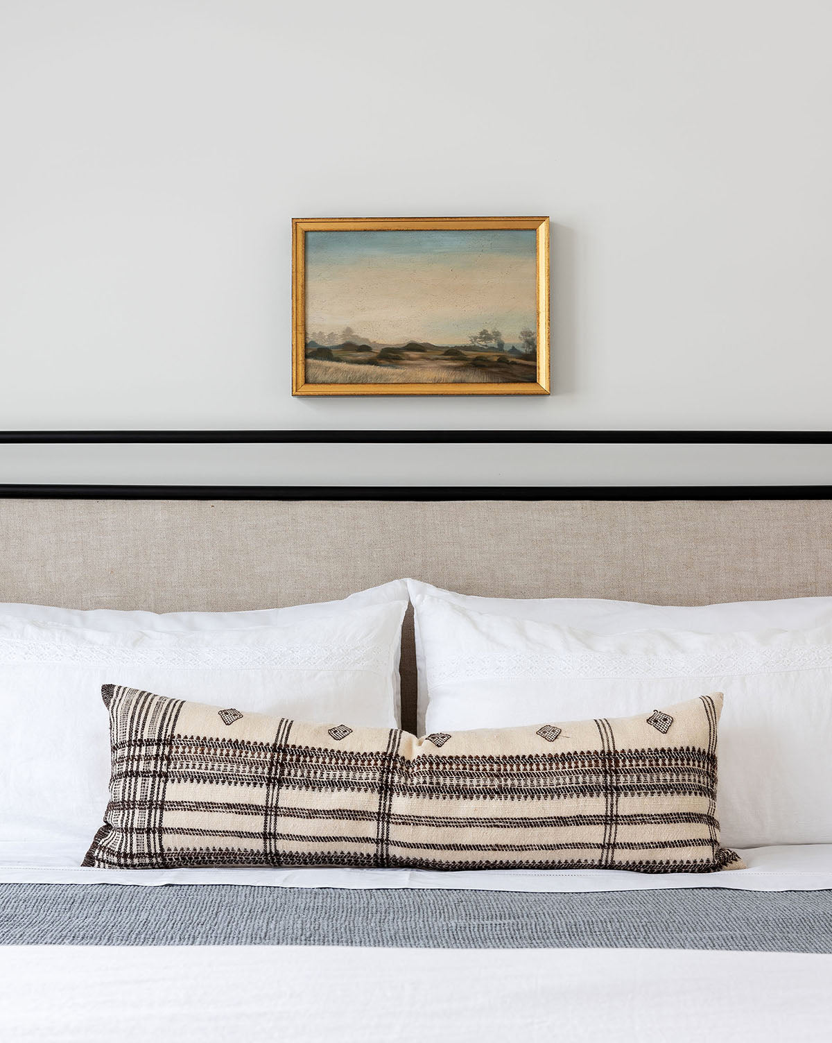 A neatly made bed with white pillows and a patterned lumbar cushion sits beneath Morning Valley, a small gold-framed landscape painting of waves of grain, displayed on a plain white wall above a light headboard.