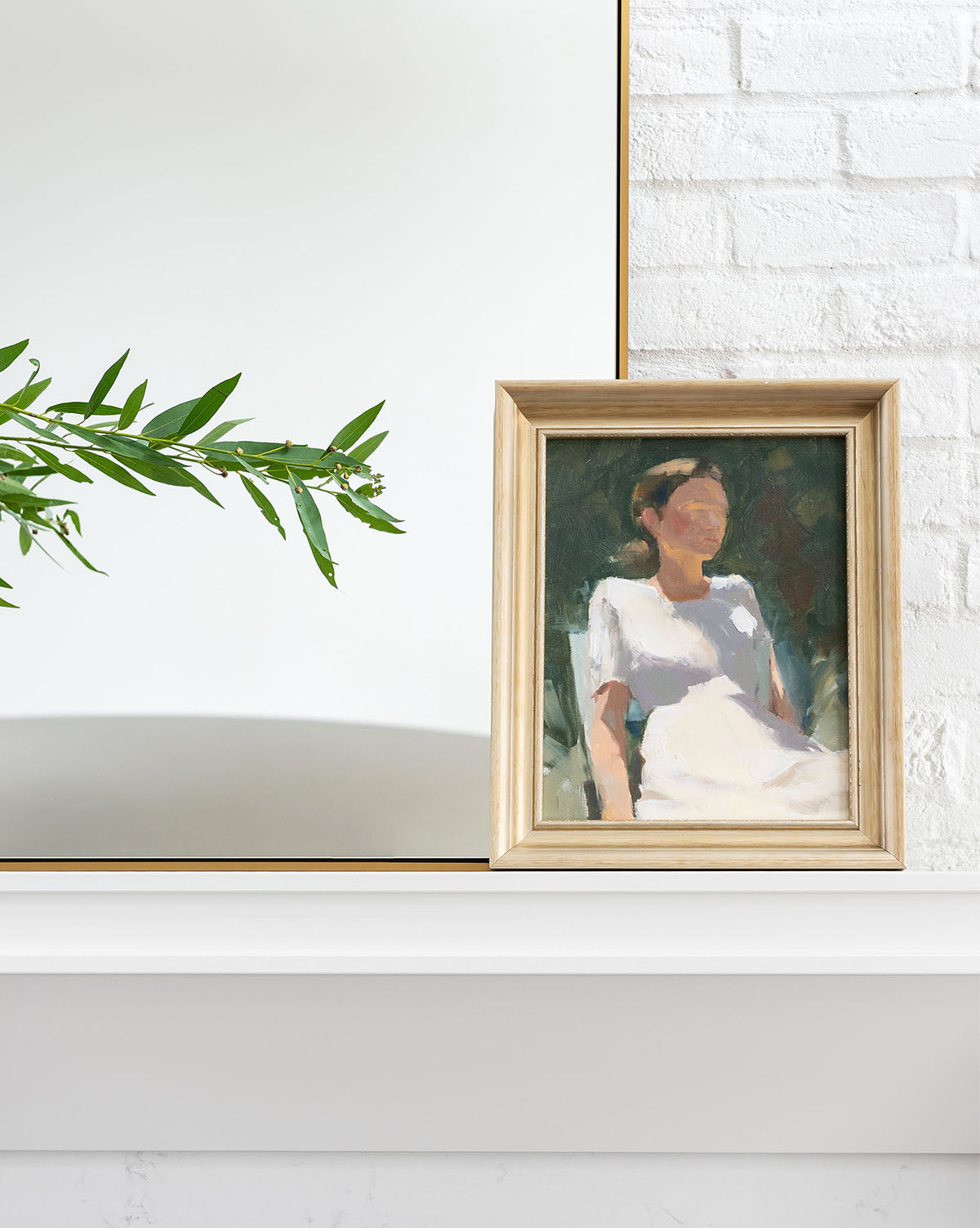 Ponder in the Park 1, a framed painting of a woman in white, sits on a white mantel before a mirror beside a leafy green branch—a serene, vintage-inspired art piece perfect for tranquil spaces.
