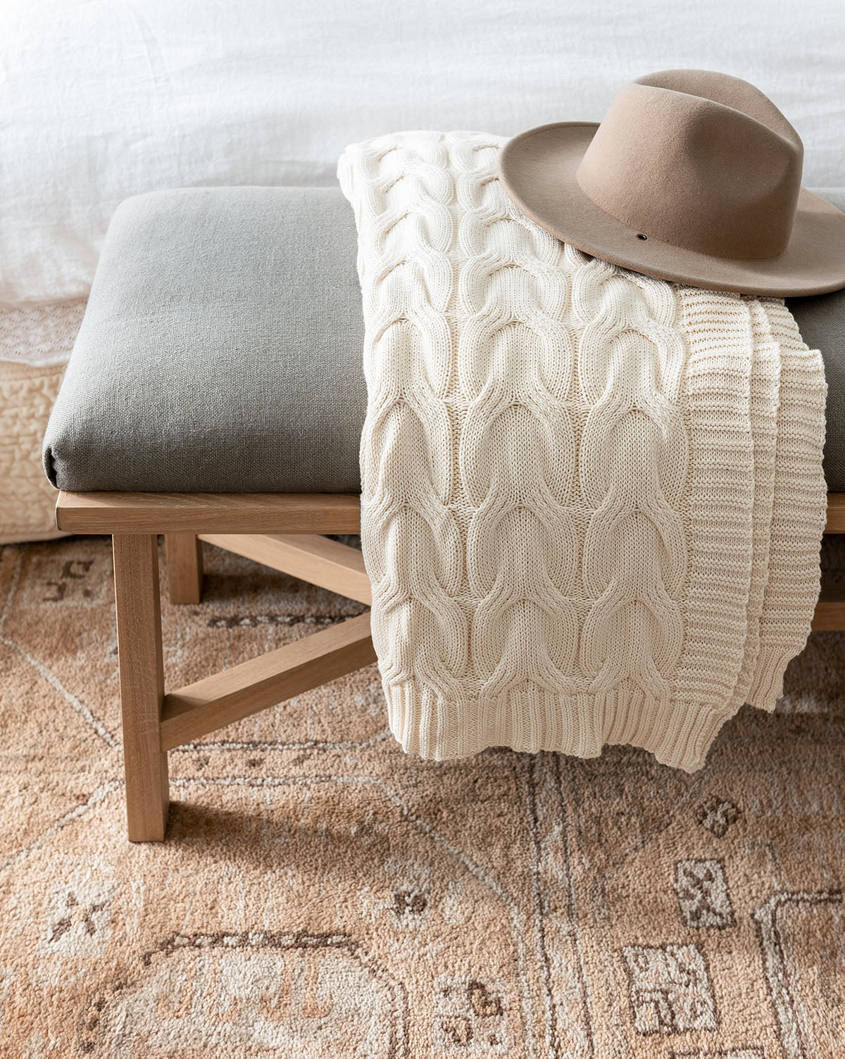 A beige knit blanket and a brown wide-brimmed hat rest on a cushioned wooden bench at the foot of a bed, atop the Ellington Fawn Hand-Tufted Wool Rug, by McGee & Co.