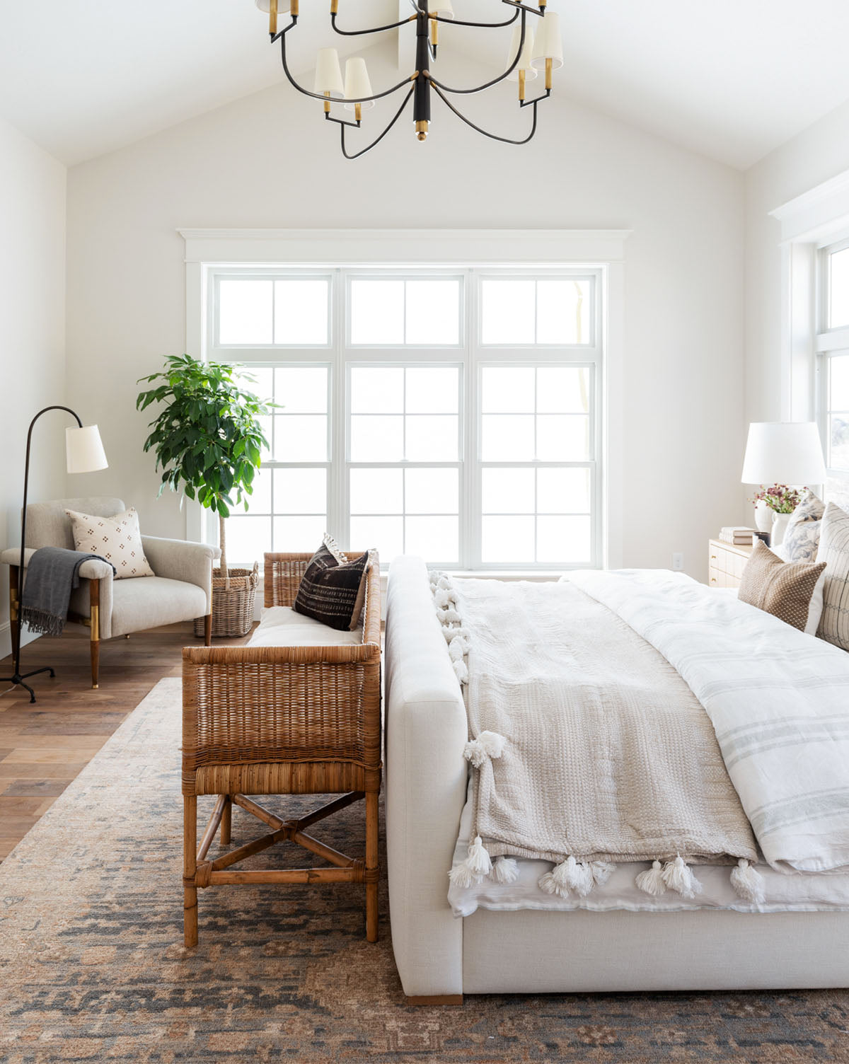 Bright, airy bedroom with large windows, a cozy armchair, potted plant, Wilshire Blue Hand-Tufted Wool Rug beside a woven bench at the bed’s foot. Light bedding, neutral decor, wood floor, and a chandelier complete the inviting space, by McGee & Co.