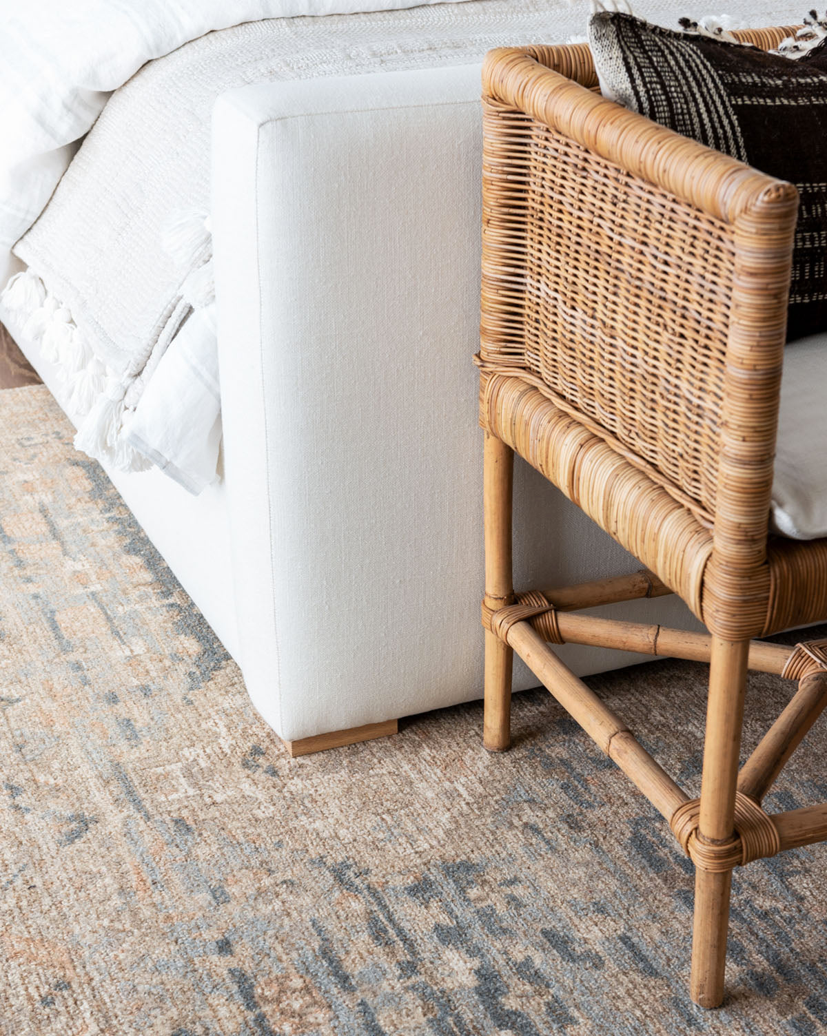 A close-up of a cozy bedroom corner featuring a white upholstered bed, a woven rattan chair with patterned pillows, and the Wilshire Blue Hand-Tufted Wool Rug, by McGee & Co.