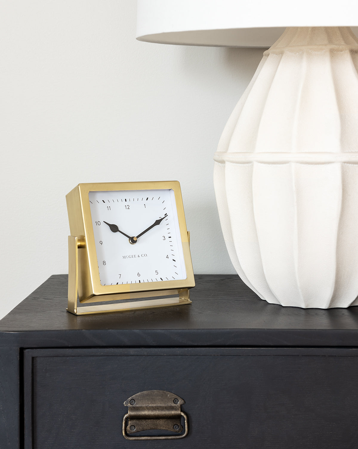 The Posey Table Clock, with its brass frame, sits on a black table beside a white textured lamp with a round base. The gold clock displays 10:09 against a light-colored background wall.