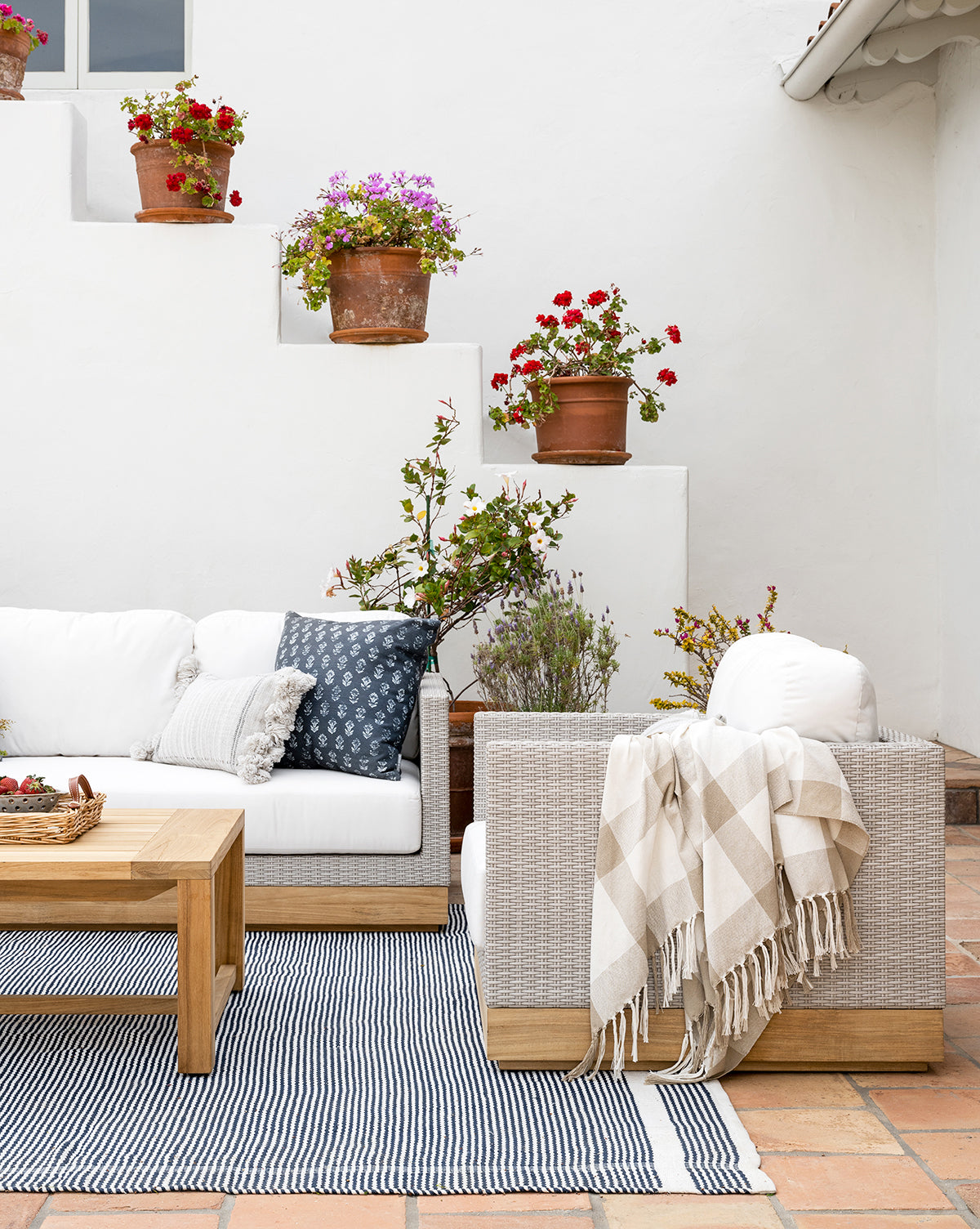 A cozy patio features the Berdine Wicker Outdoor Chair with white cushions, throw pillows, and a blanket. A wooden coffee table and striped rug sit on terracotta tiles. Potted flowers decorate stair-like shelves against a white wall.