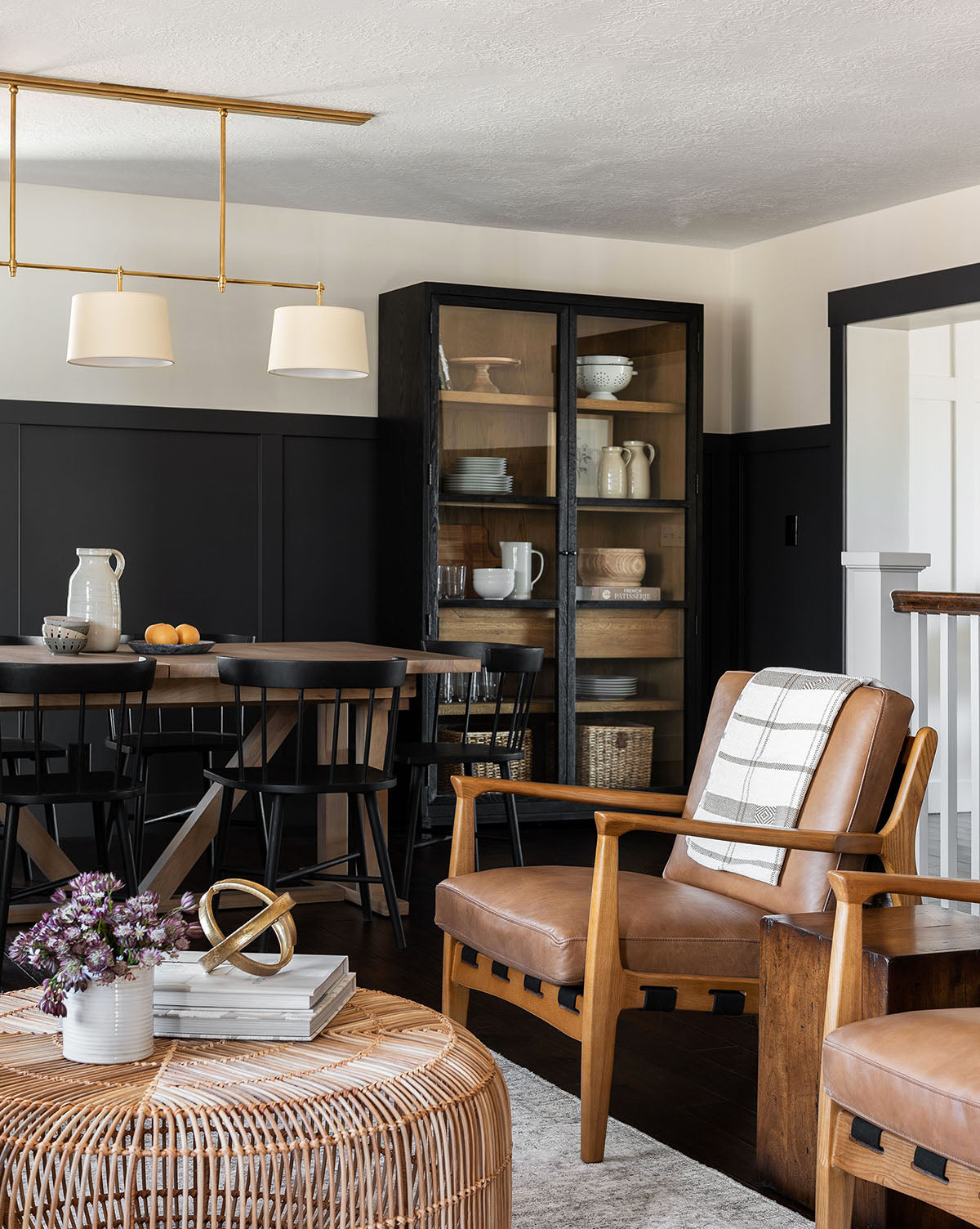 A modern dining and living space features black wainscoting, a wooden dining table, Lanston Lounge Chairs, a glass-door cabinet, leather armchairs with ash wood frames, a round rattan coffee table with books and flowers, and a white pendant light.