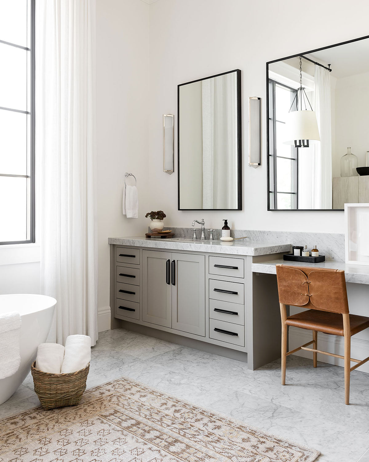 A modern bathroom with a freestanding tub, gray vanity with black handles, double mirrors, wall sconces, marble countertop, Danson Dining Chair with leather seat, towels in a basket, and a patterned rug on marble flooring by McGee & Co.
