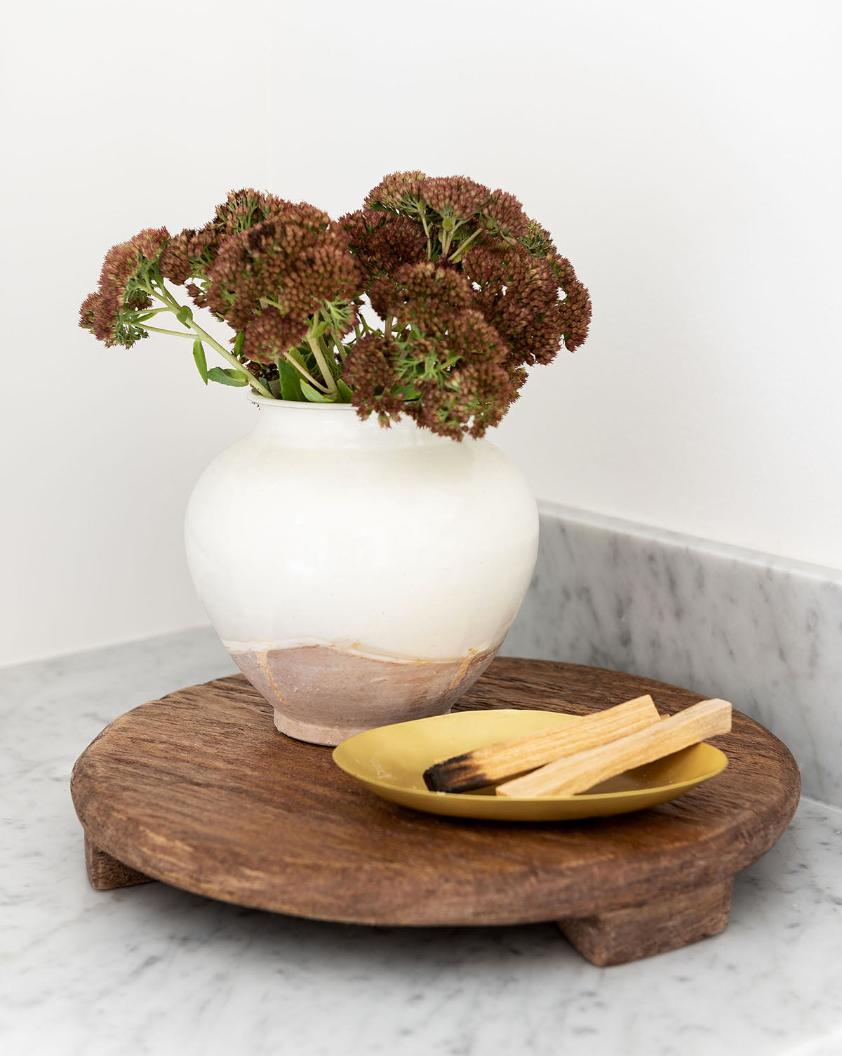 A white ceramic vase with dried brown flowers is displayed on a Mango Wood Carved Pedestal beside a gold dish of palo santo sticks, all arranged on a marble countertop.