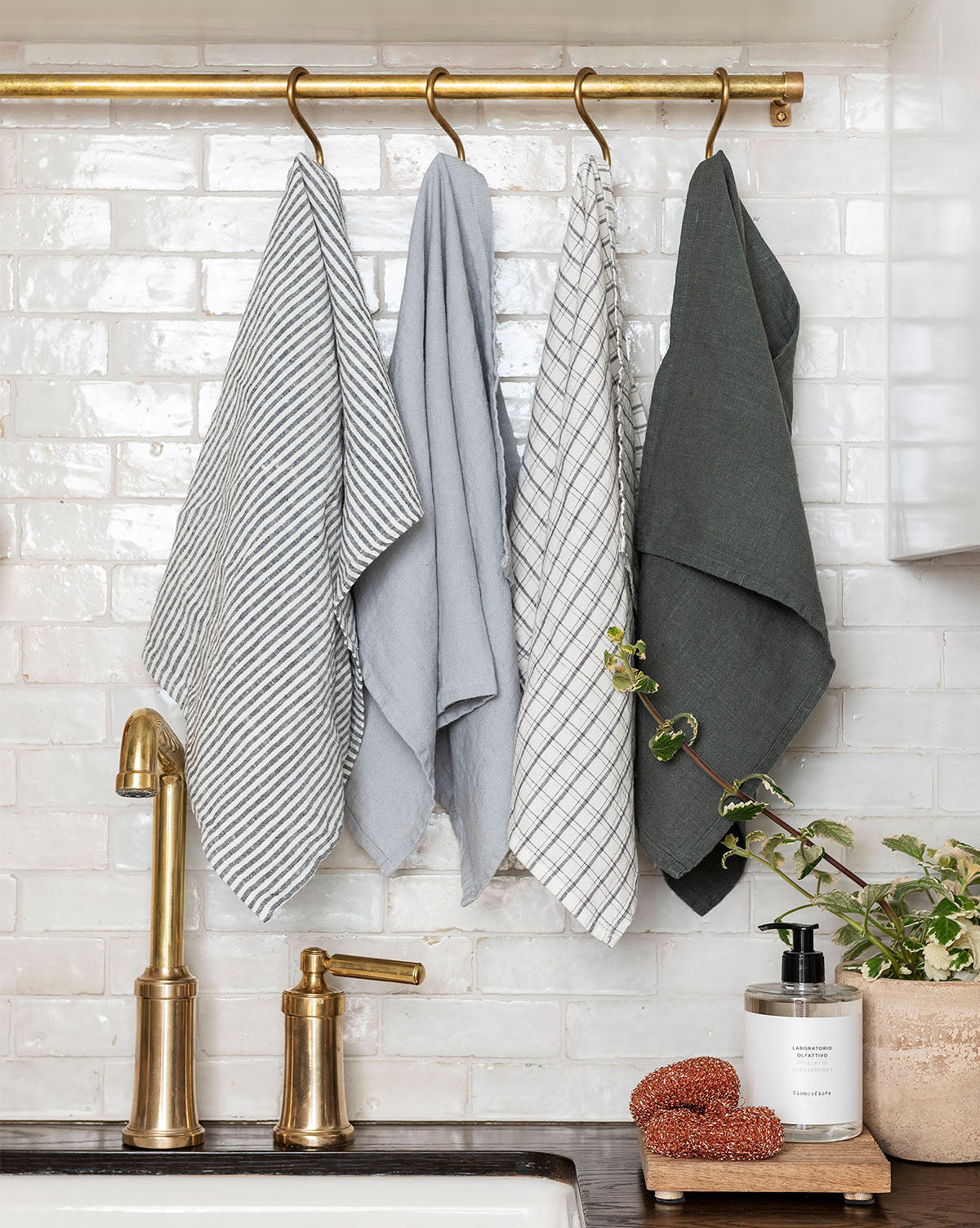 Four kitchen towels hang from a brass rod above a sink with a gold faucet. Laboratorio Olfattivo Liquid Soap, a plant, and scrubbers sit on a wooden tray on the countertop. White tile forms the backsplash.