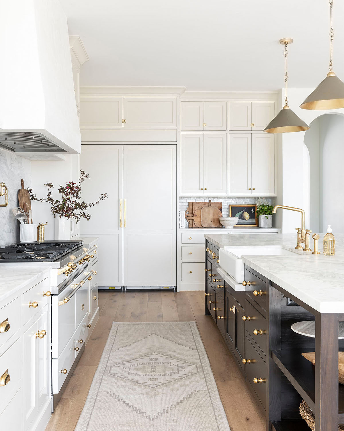 Bright, elegant kitchen with white cabinets, gold hardware, marble countertops, a farmhouse sink, pendant lights, and the Pembroke Hand-Knotted Wool Rug on light wood floors. Decorative plants and dishes adorn the counters and shelves, by McGee & Co.