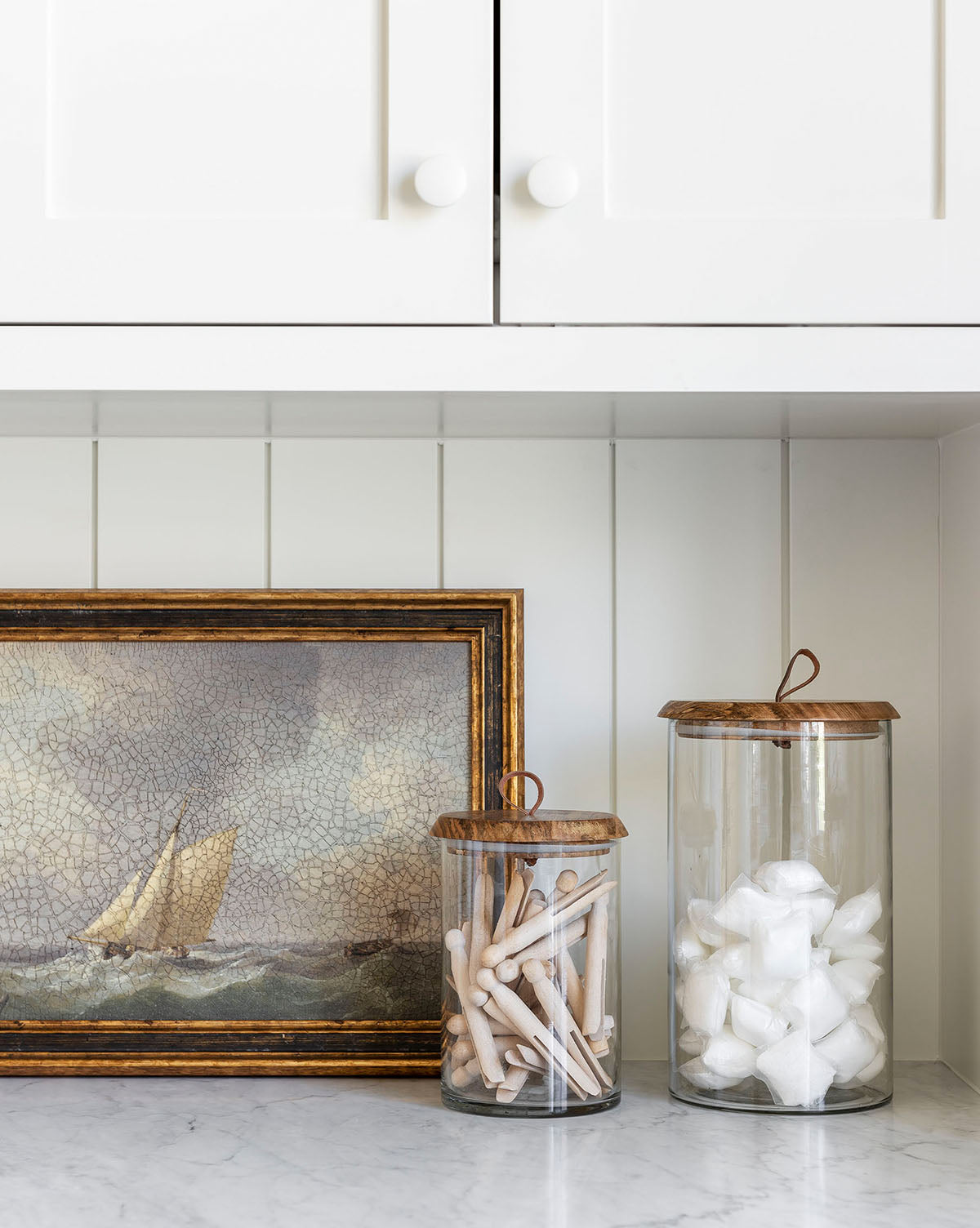Two Seafarer glass jars filled with cotton swabs and cotton balls sit on a white countertop beside a sailboat painting, adding subtle coastal charm to the white paneled wall and cabinet.