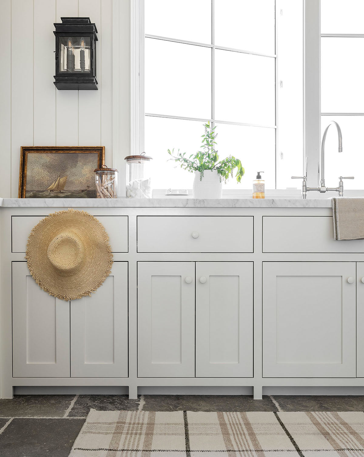 The Seafarer kitchen features white cabinets, a marble countertop, a large window, potted greenery, vintage accents, a straw hat on a drawer, a framed sailboat painting for nautical charm, and a plaid rug on the stone floor.