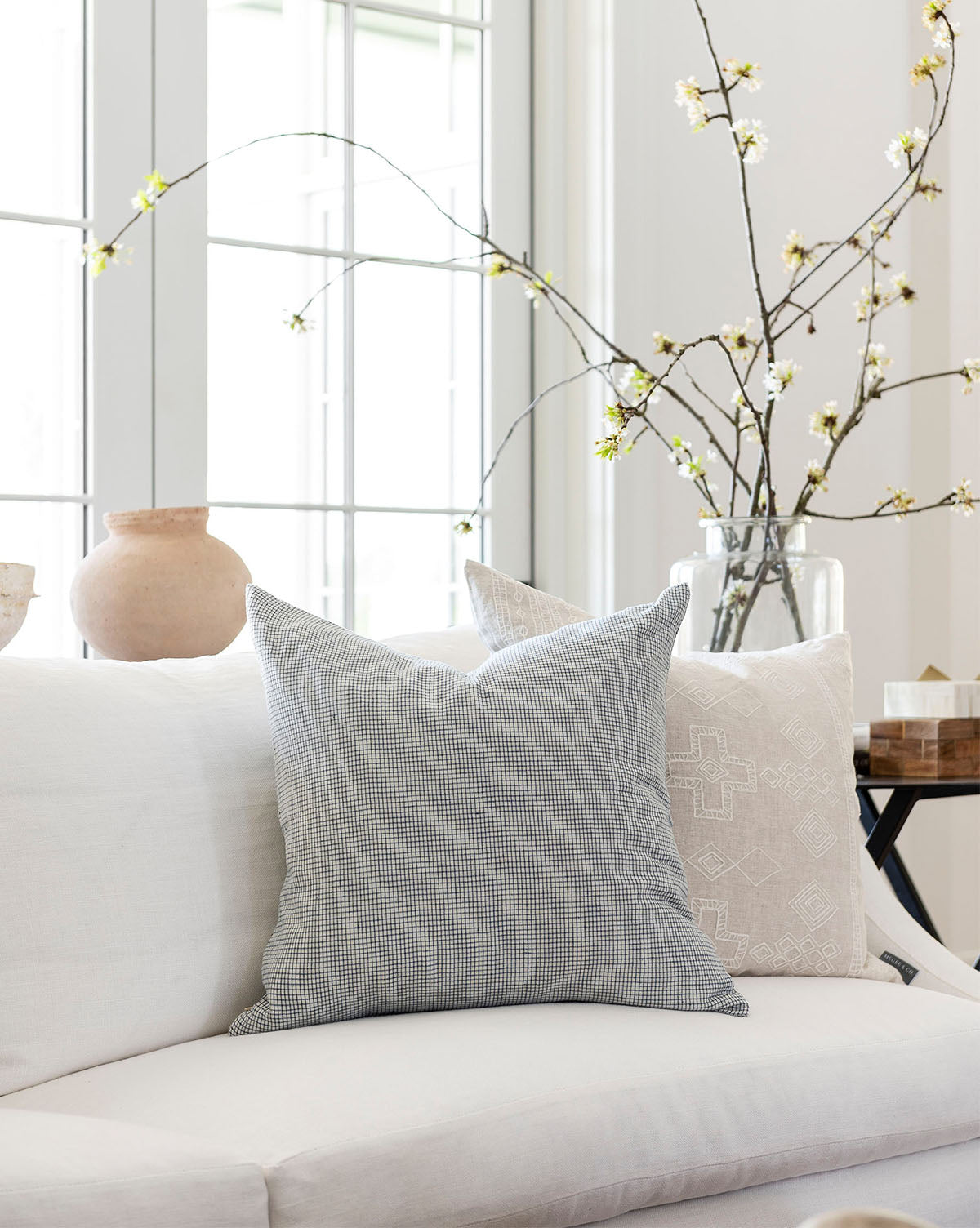 A cozy white sofa with two decorative pillows, including the Luther Pillow Cover in blue and cream, sits in a bright room. Nearby, a large glass vase with blossoming branches adds a touch of nature by tall sunlit windows, by McGee & Co.