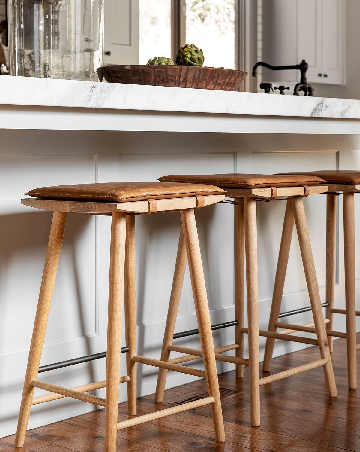 Three Beau Counter Stools with brown wooden seats and a minimalist design are lined up at a white marble kitchen island, adding a stylish touch, by McGee & Co. Part of the kitchen counter and decor can be seen in the background.