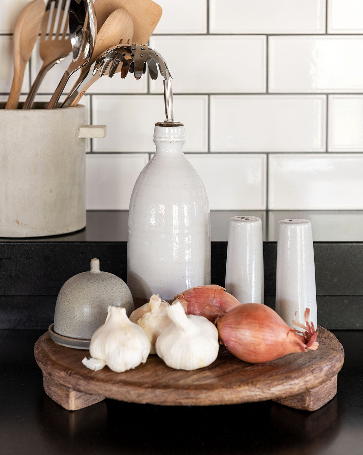 The Mango Wood Carved Pedestal displays a wooden tray with garlic, shallots, condiments, and shakers on a black kitchen counter featuring a utensil holder and white subway tile backsplash.