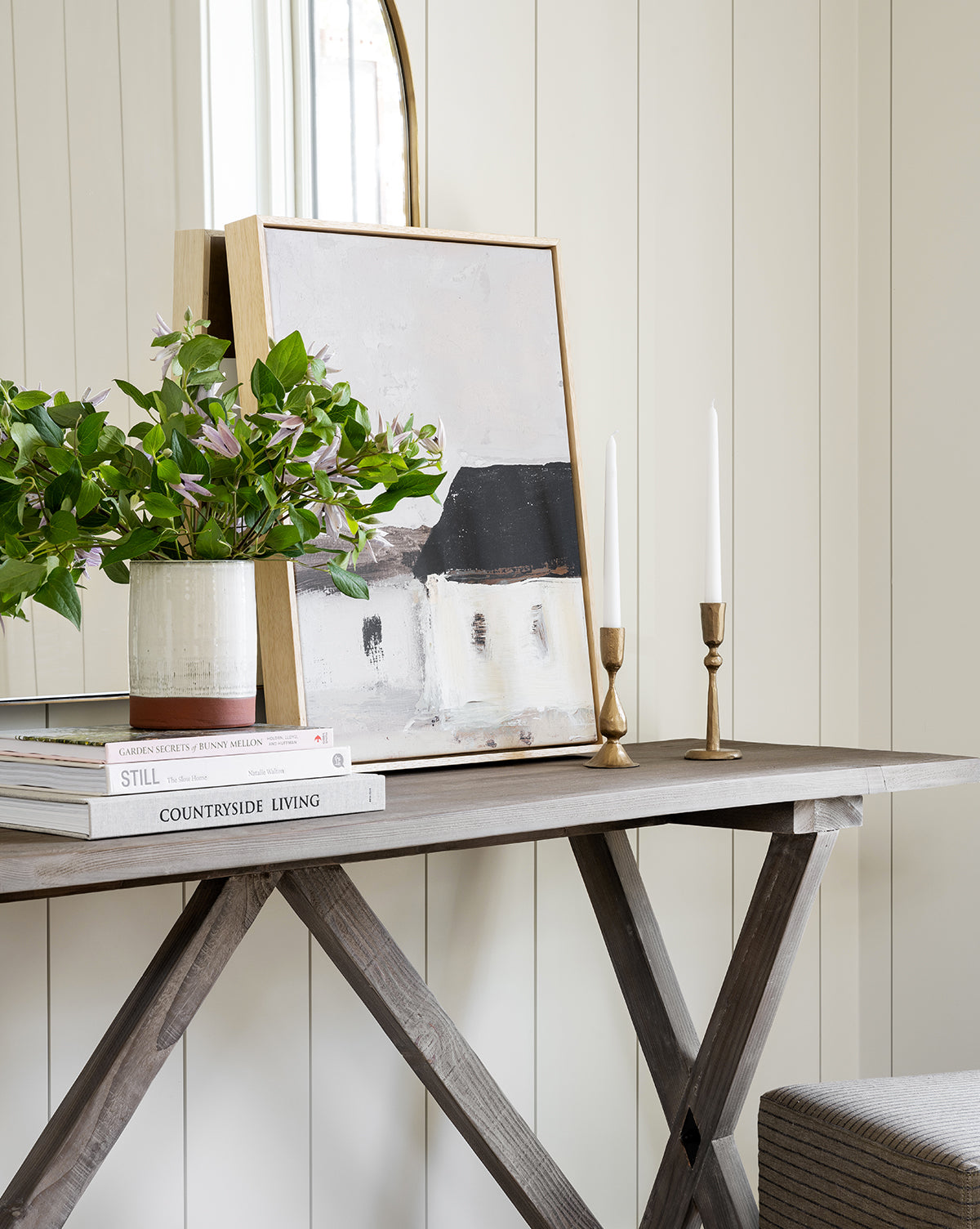 A wooden console table topped with a potted plant, stacked books, two brass candlesticks, and the Hidden Barn by Donna Thomas artwork creates a cozy vignette against the cream-paneled wall.