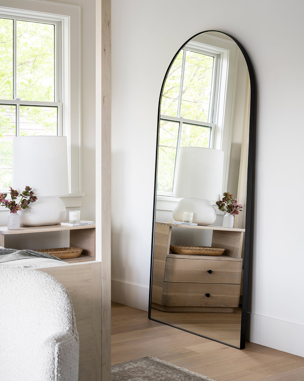 The Gala Floor Mirror with a black frame is positioned against a white wall, reflecting a window, wooden nightstand with drawers, white lamp, and flower vase in this bright modern bedroom by McGee & Co.