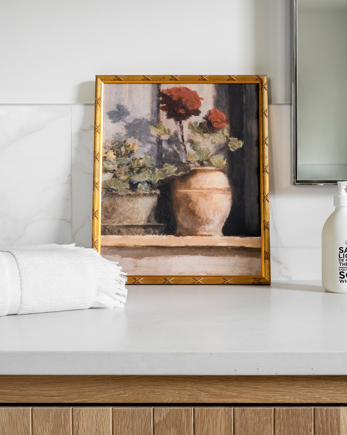 A framed Stoop Geraniums painting sits on a white countertop near a folded towel and soap dispenser in a modern bathroom with light wood, marble accents, and soft summer light.