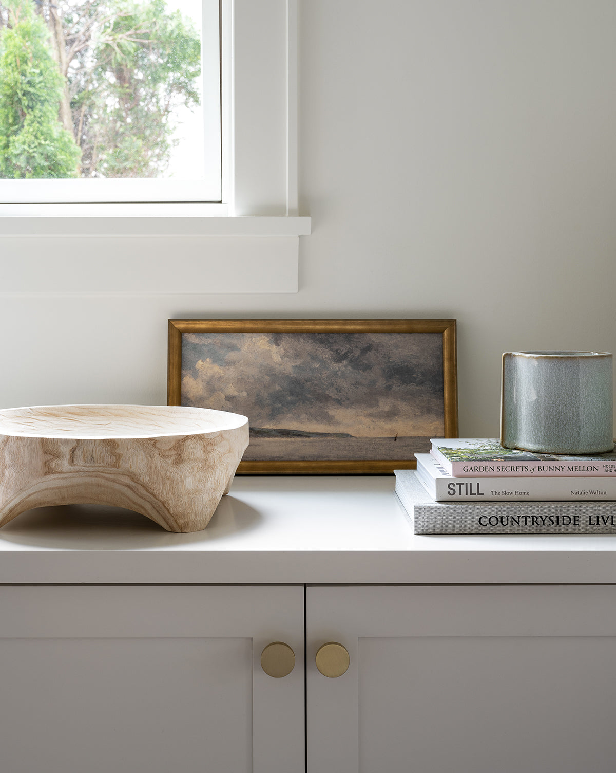 A white cabinet with gold knobs holds a wooden bowl, stacked books, a blue-green candle, and Gray Seas, a framed gray-toned artwork of clouds, under a window with greenery visible outside.