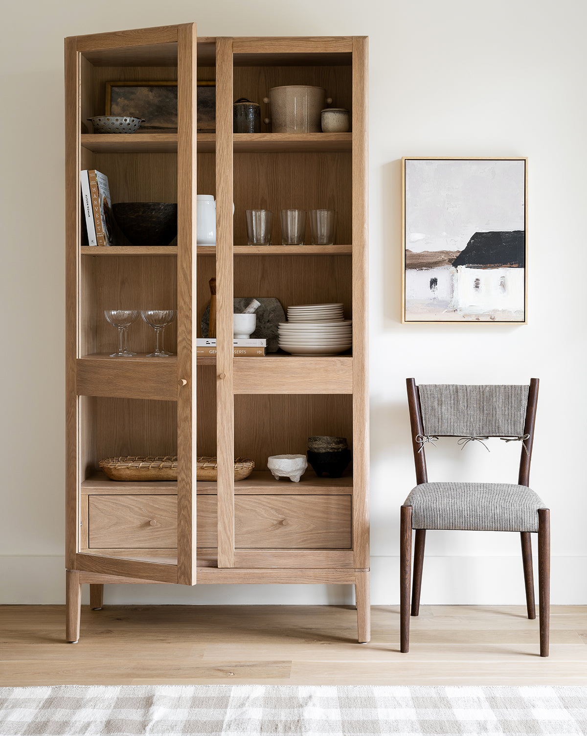 A gray cushioned chair sits beside a wooden cabinet with glass doors displaying dishes, under Hidden Barn by Donna Thomas, perfect for gallery wall lovers. A light-striped rug complements the white wall and wooden floor.
