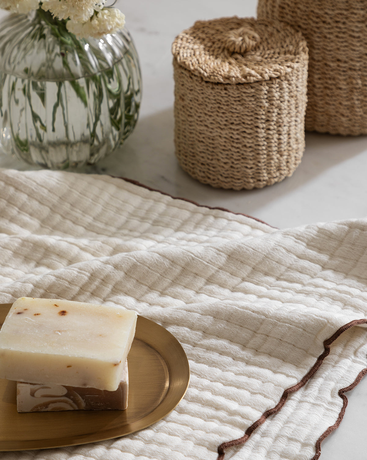 Two soap bars sit on a brass tray atop the Cassidy Waffle Hand Towel, a textured cream cotton towel with brown trim. Behind them are two woven containers and a glass vase with white flowers.