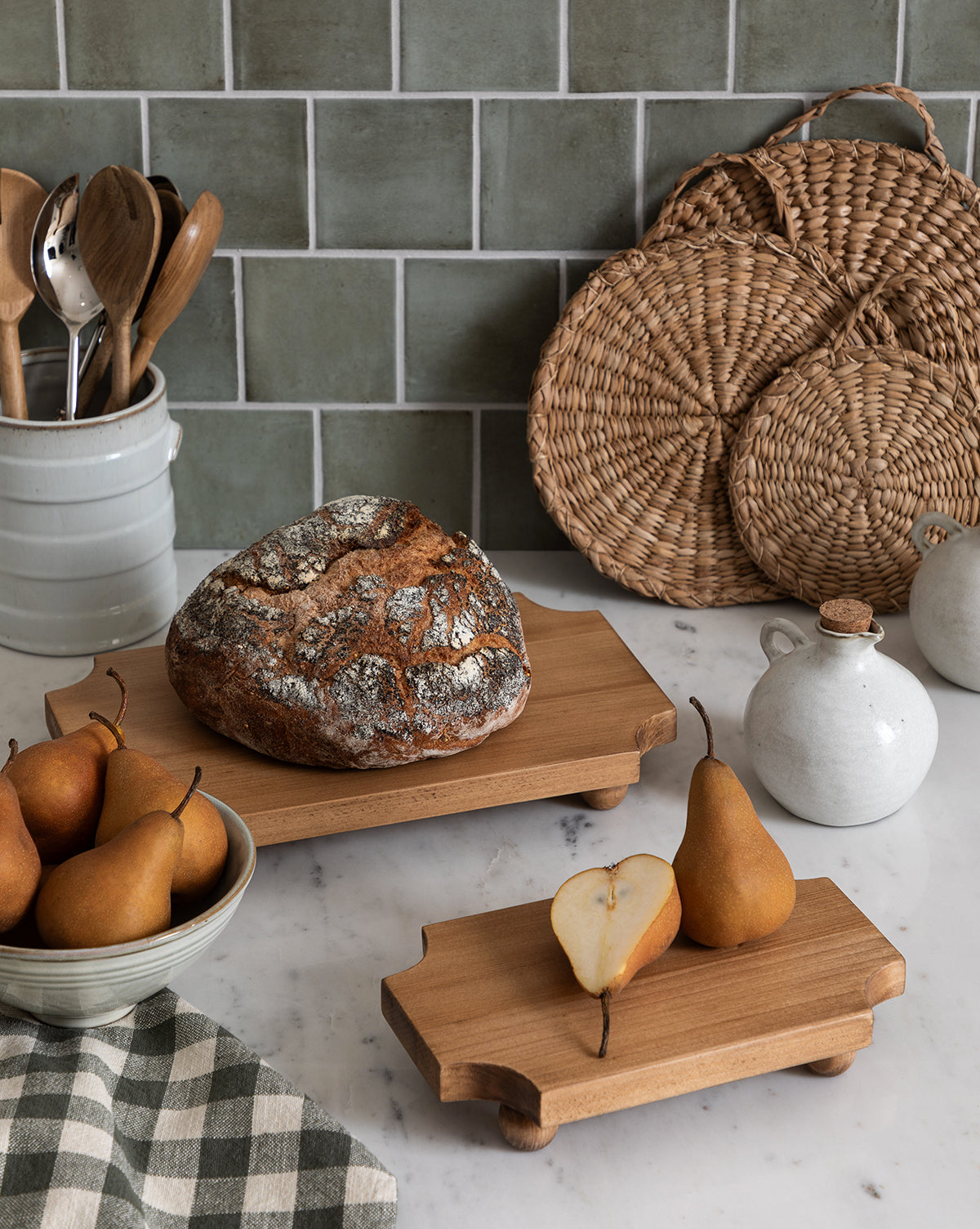 A rustic kitchen scene with pears in a bowl, a sliced pear on the Cillian Pedestal Board, a round loaf of bread, ceramic jars, wooden utensils in a crock, woven placemats, and a checkered cloth on a marble countertop by McGee & Co.