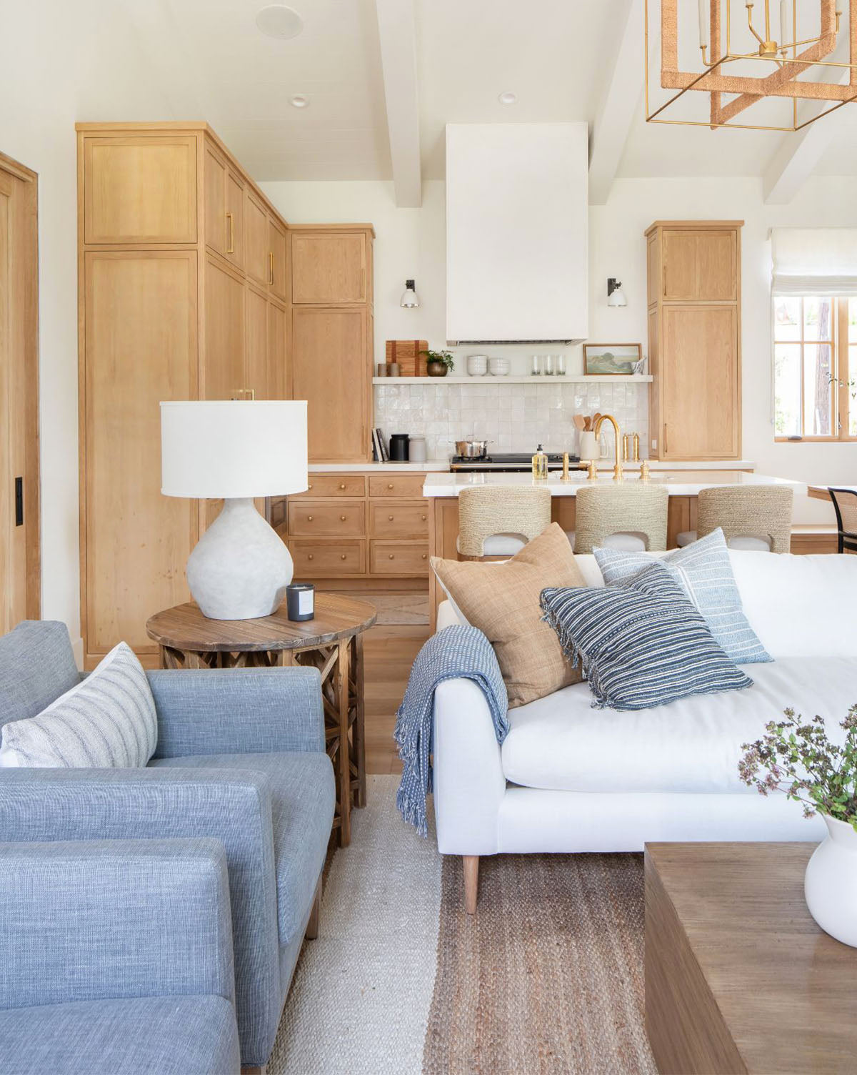 Bright, modern living room and kitchen with light wood cabinets, a white sofa accented by blue and tan pillows, gray armchair, neutral rug, potted plant on a wooden coffee table, and the Stetson Table Lamp on the side table, by McGee & Co.