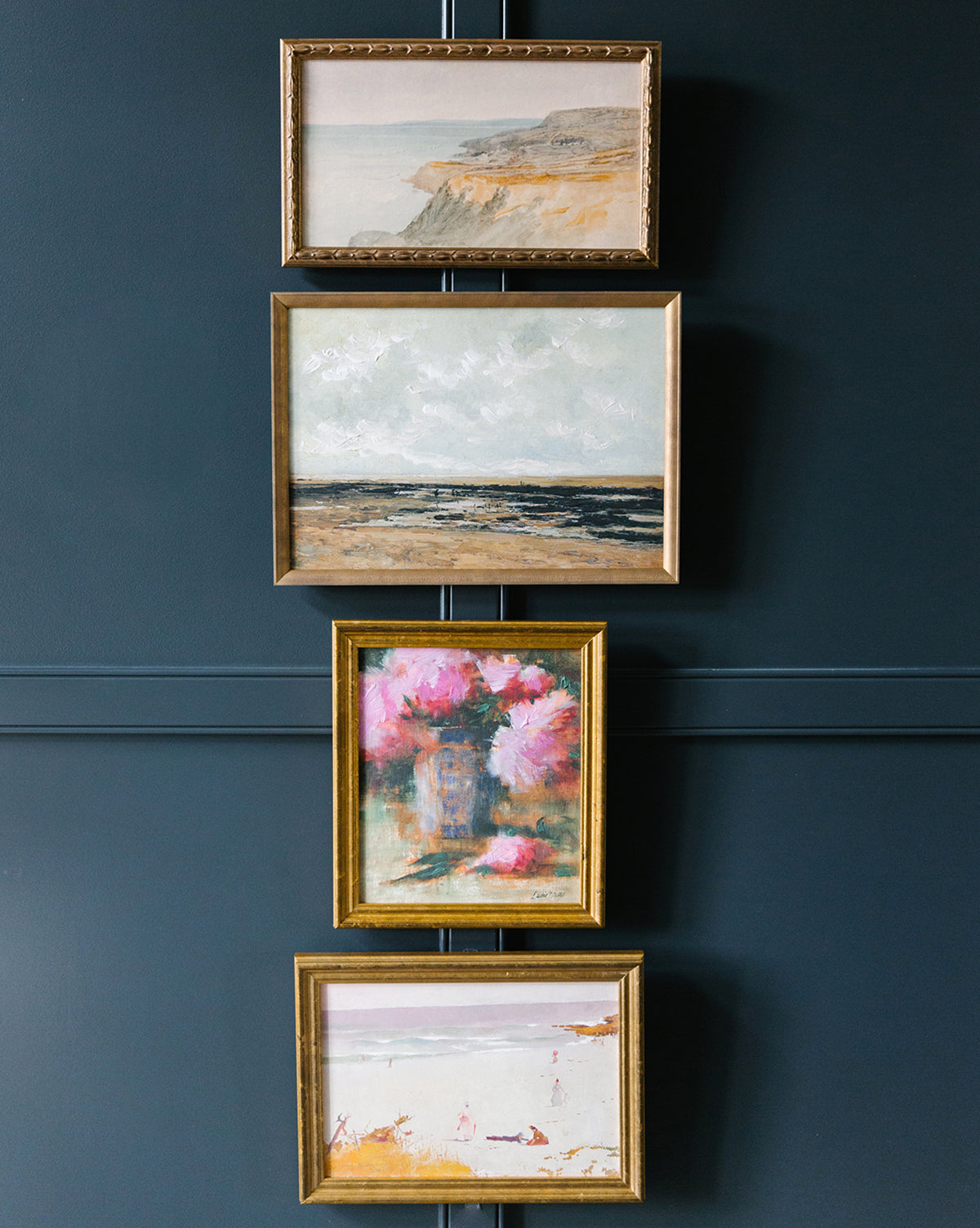 Four framed paintings hang vertically on a dark blue wall, featuring a vintage cliffside, an ocean view, the Peonies & Vase still life with pink petals, and a beach scene with people.