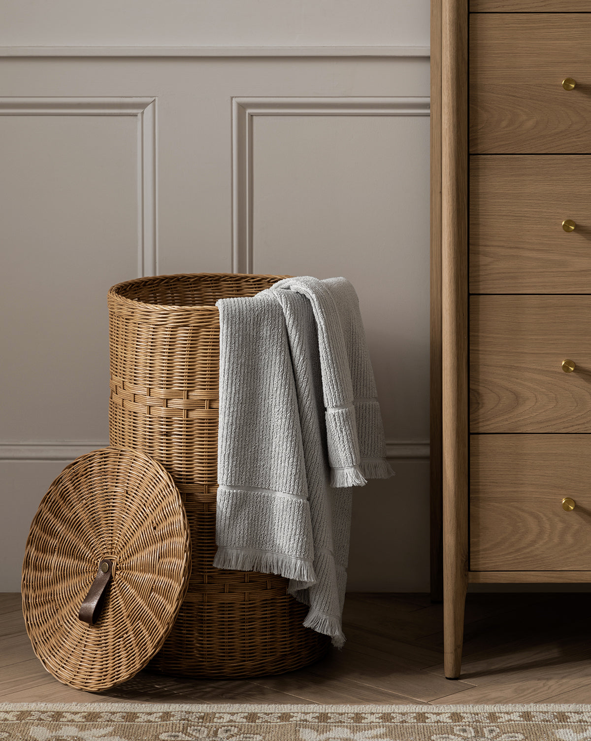 A wicker laundry basket with its lid off, draped with a folded towel from the Edessa Bath Collection, sits beside a wooden dresser in a paneled room with a patterned rug.