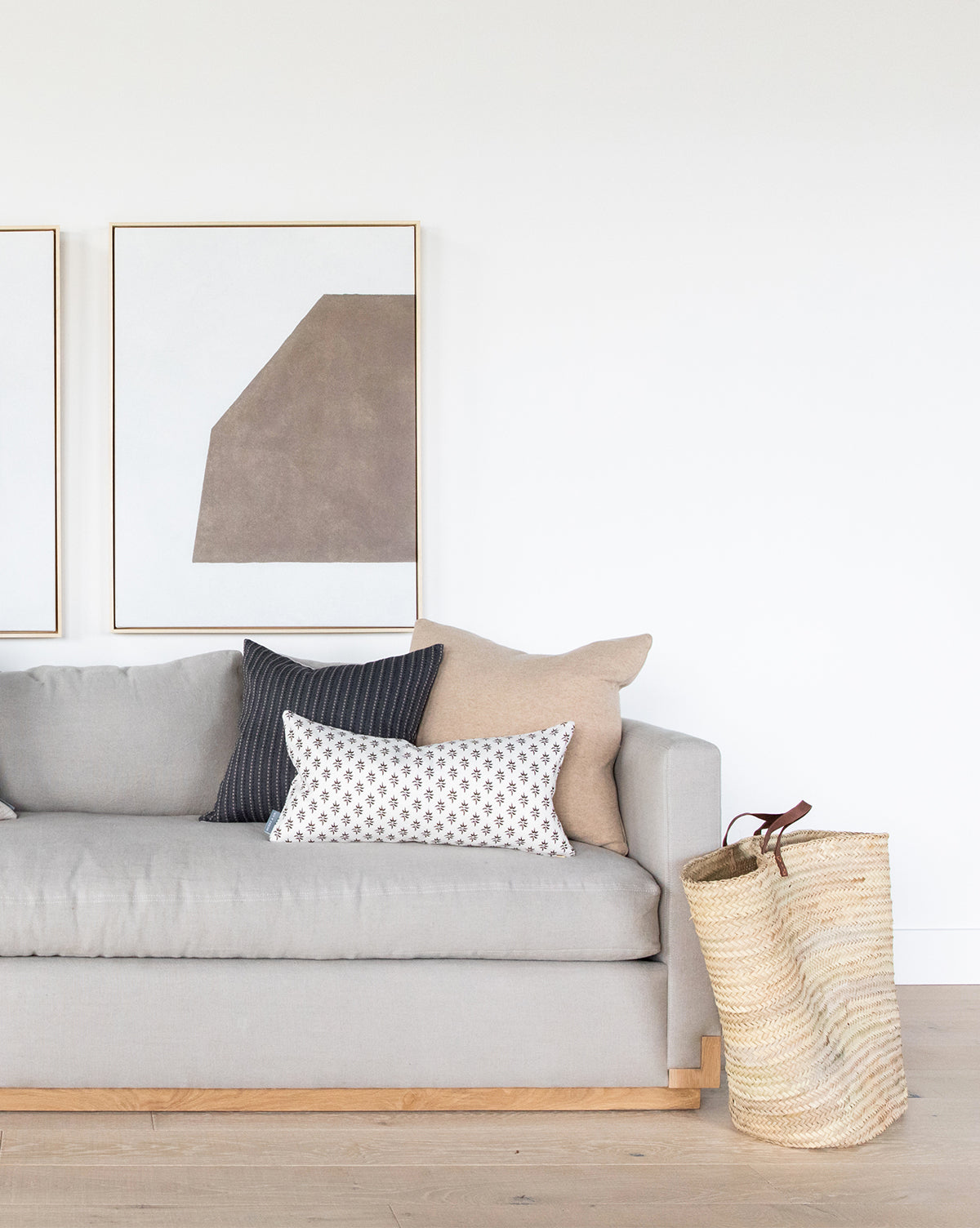 A modern living room with a light gray sofa, neutral throw pillows, a woven basket on the floor, and curated wall art featuring the Entering Shape 2 abstract framed piece on a white wall in the background.