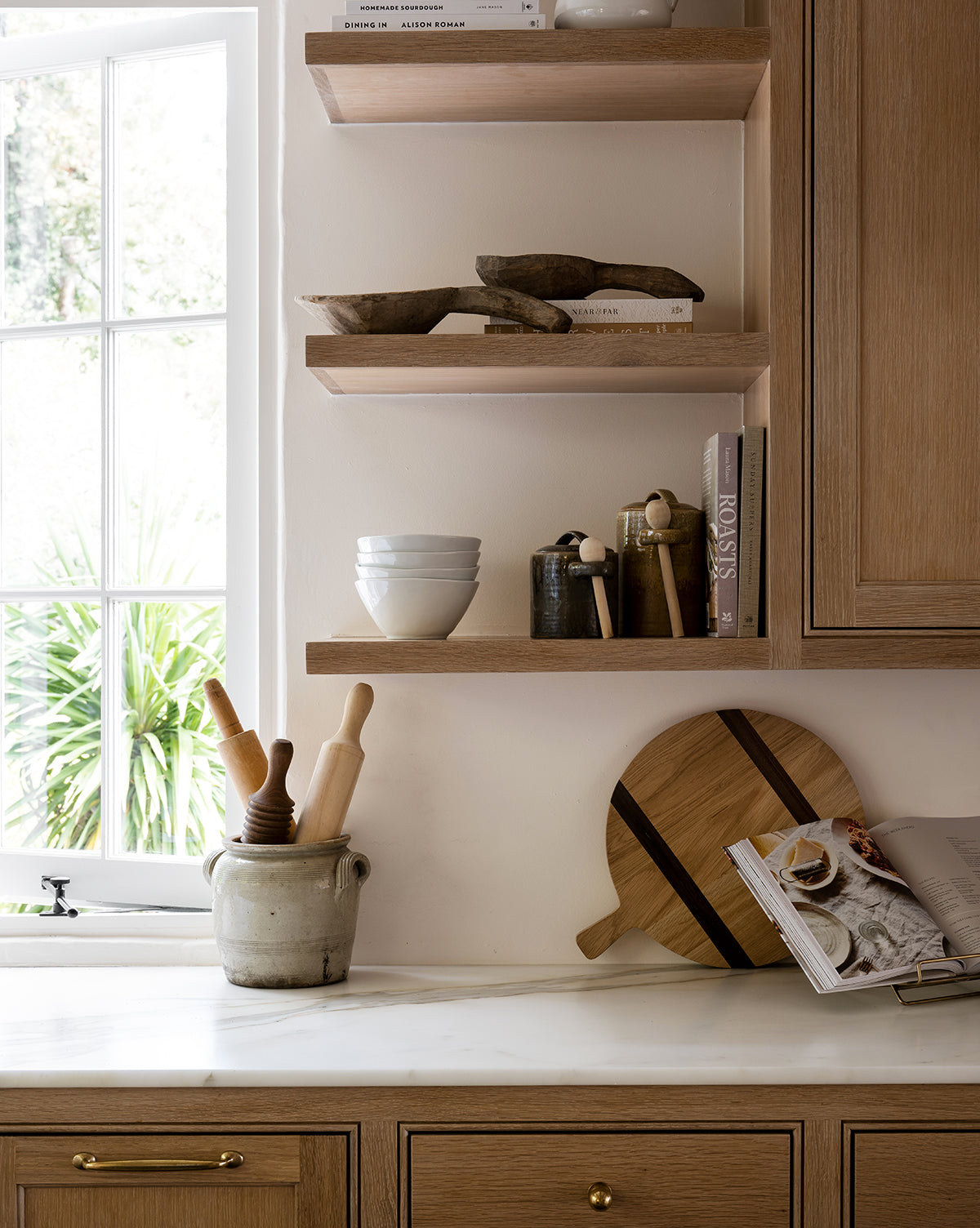 A bright kitchen corner with wooden shelves displays bowls, books, and utensils. Sunlight highlights a marble counter set with a crock of rolling pins, cutting boards like the Round Oak Bread Board, a pepper mill, and an open cookbook by McGee & Co.