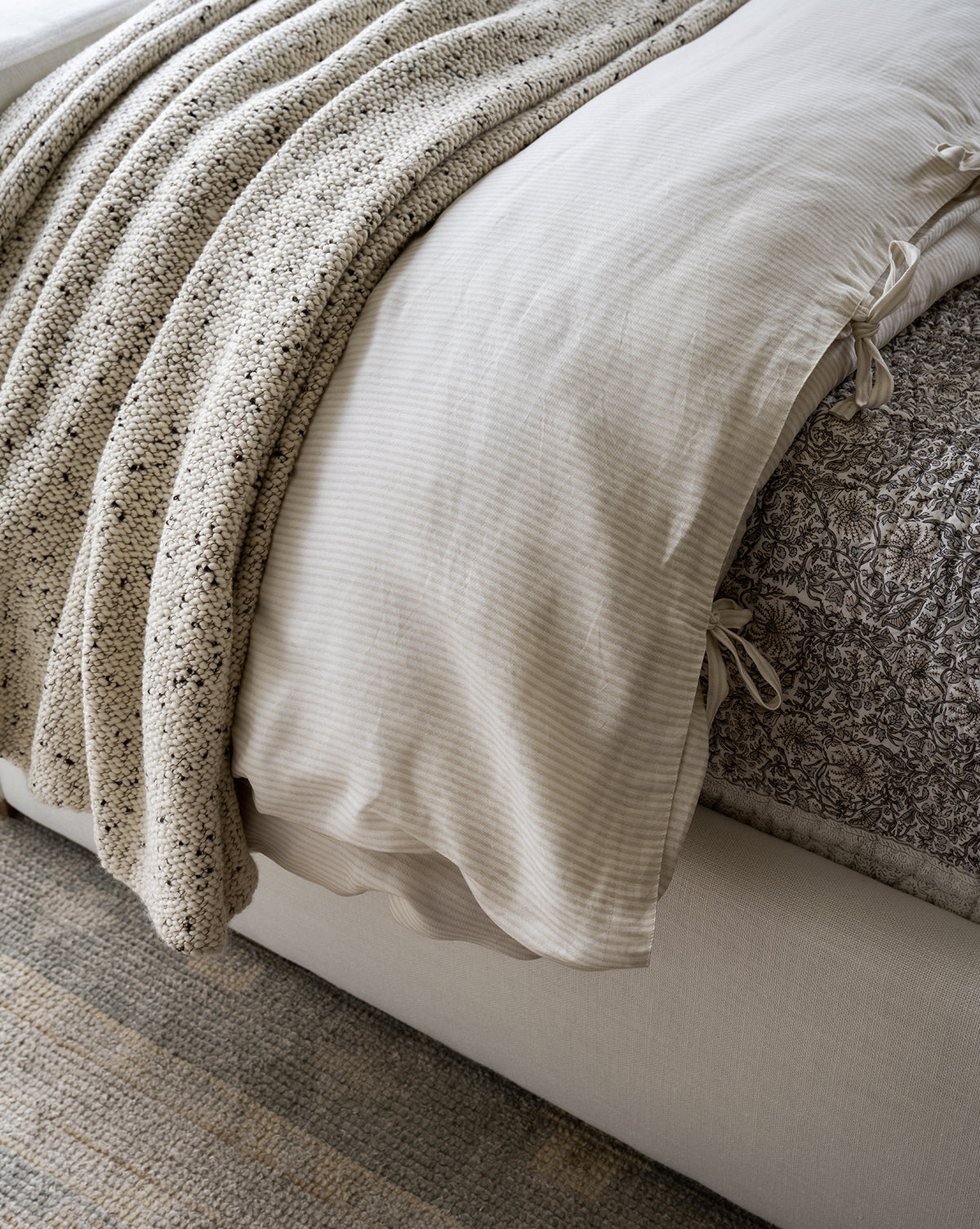 A close-up of a neatly made bed featuring the elegant Easton Striped Duvet Cover, a textured beige knit blanket, and a patterned gray sheet on a fabric-covered bed frame.