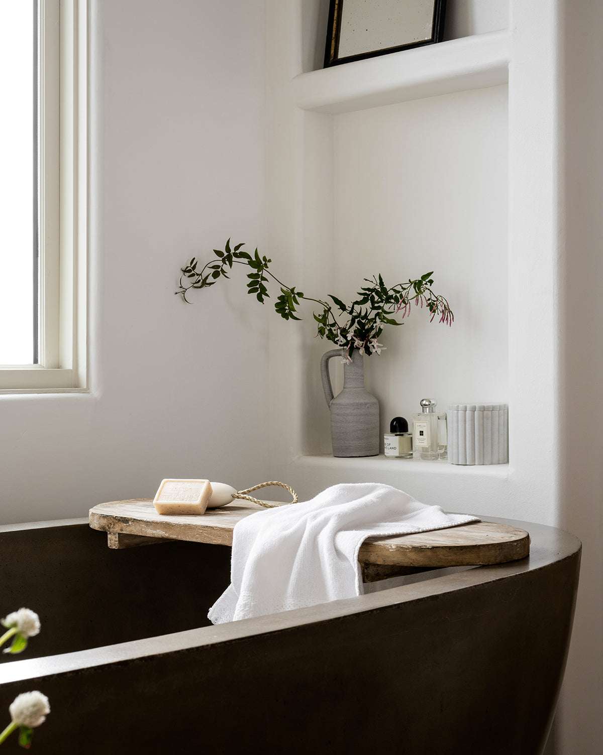 A modern bathroom with a dark bathtub features a Rounded Edge Bath Caddy holding a white towel and soap, built-in shelves with a vase of greenery, toiletries, and rolled towels. Sunlight streams in through the nearby window.