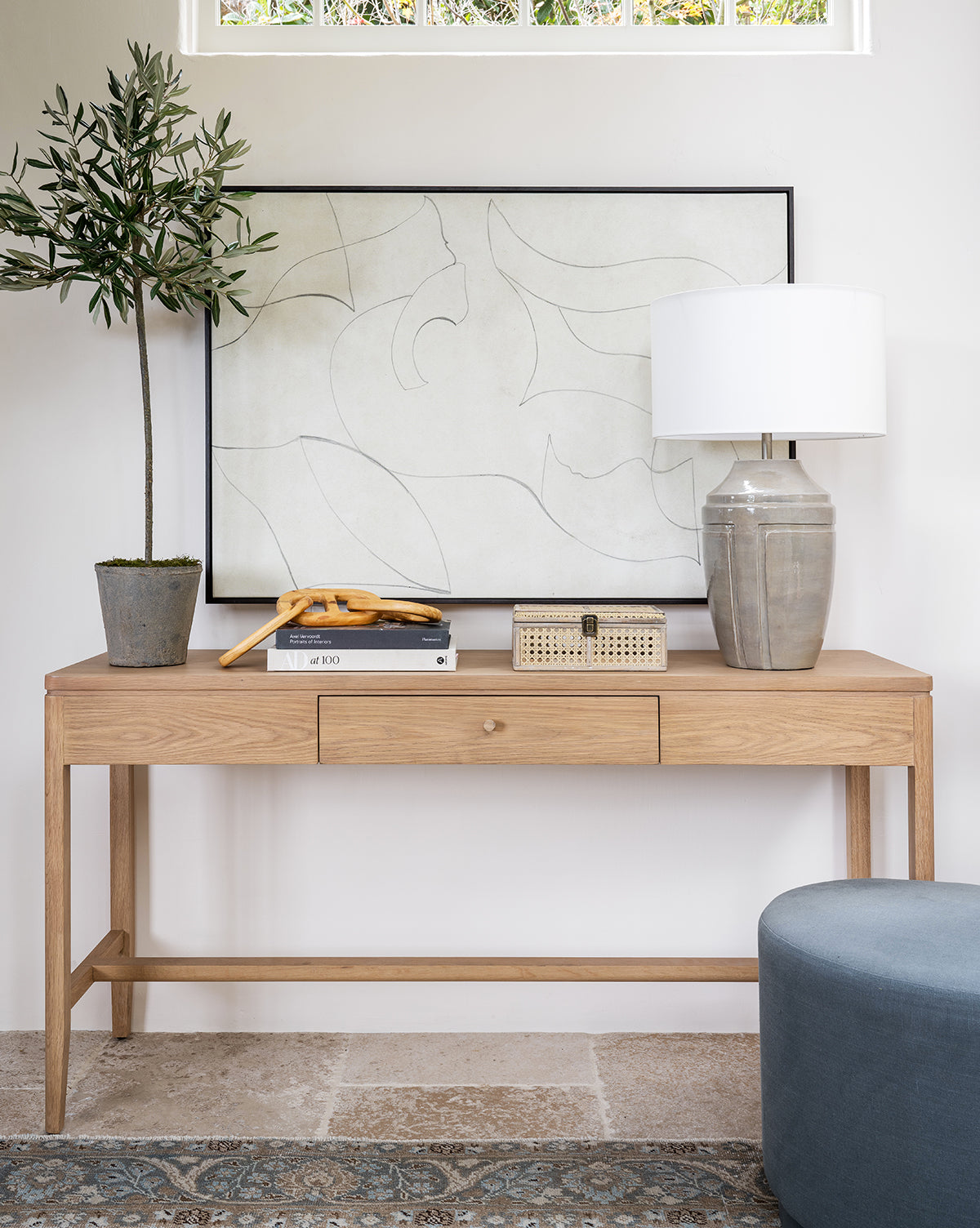 A wooden console table displays a potted plant, bananas on a plate, a decorative box, a book, and a gray ceramic lamp with white shade; Sketched Thoughts abstract canvas art hangs on the wall behind.
