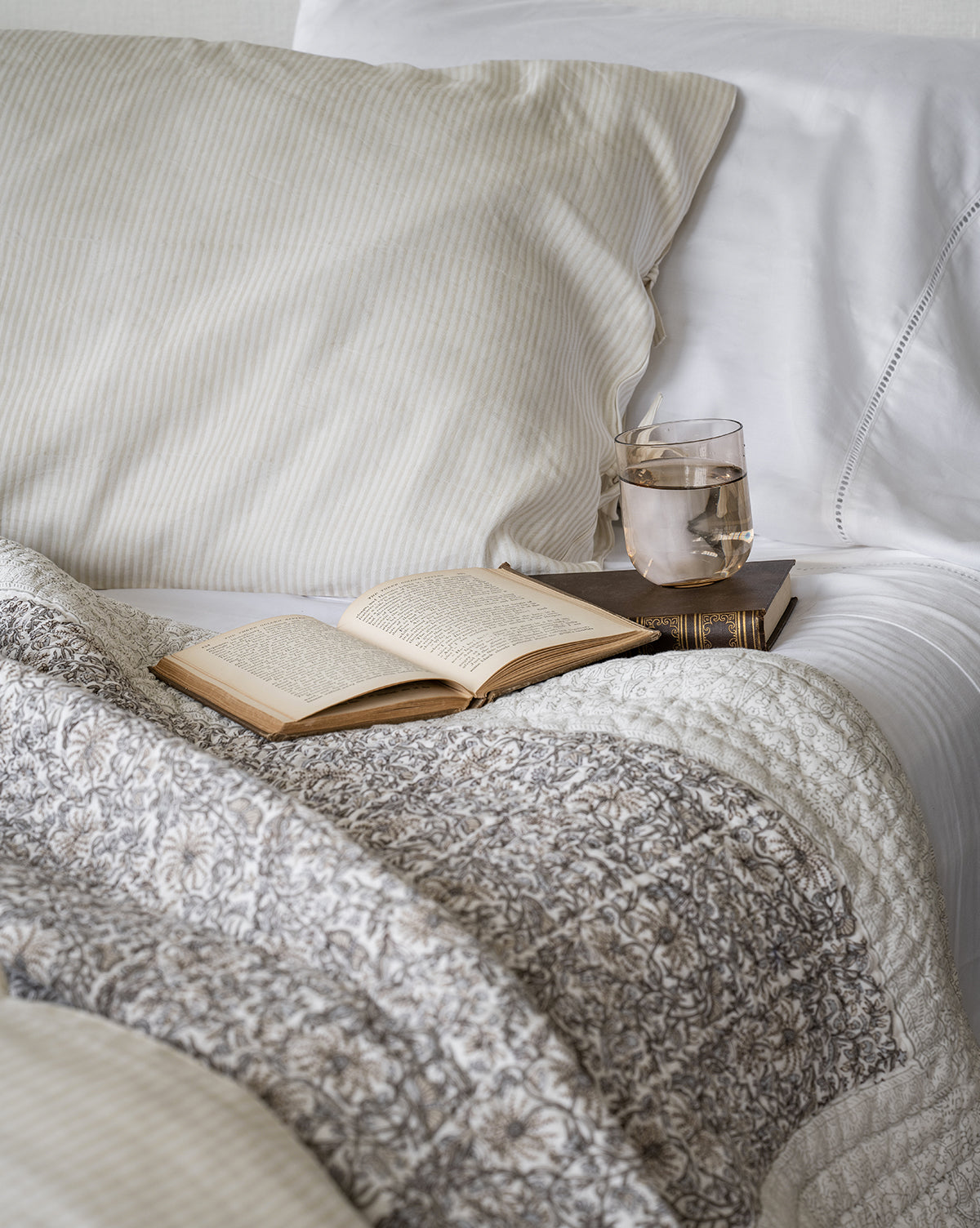 An open book, a closed book, and a glass of water rest on a bed adorned with the Floral Block Print Quilt and striped pillows.