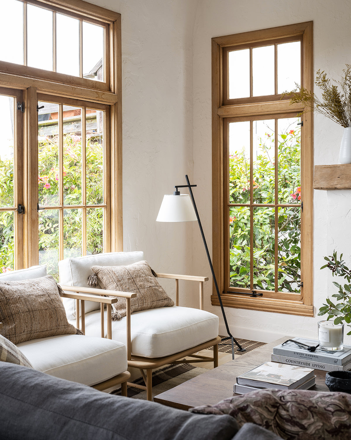 Bright living room with large windows, wooden frames, an Alma Lounge Chair with plush cushions, two cushioned chairs with textured pillows, a modern floor lamp, coffee table stacked with books, and greenery outside. The space feels cozy and inviting by McGee & Co.