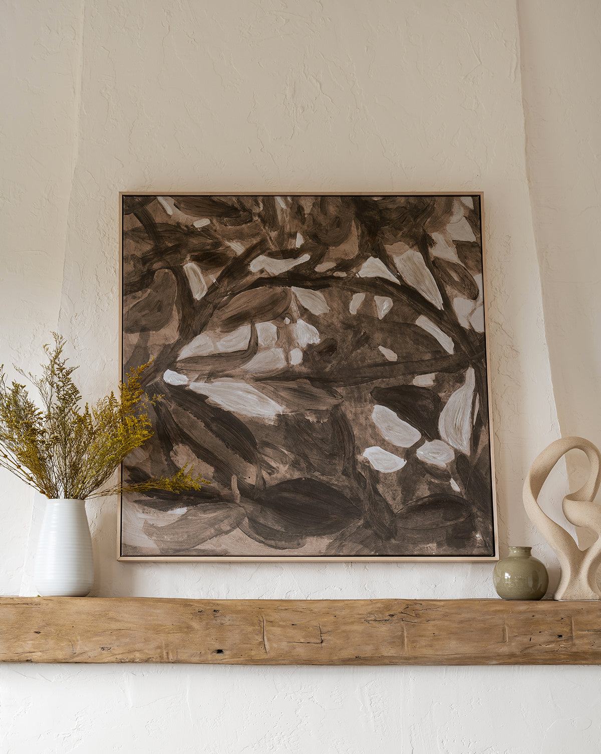 Light of Summer by Cheryl Clinton, a framed abstract wall art in brown, black, and white tones, hangs on a textured white wall above a rustic wooden shelf styled with a white vase of yellow branches, green bowl, and beige abstract sculpture.