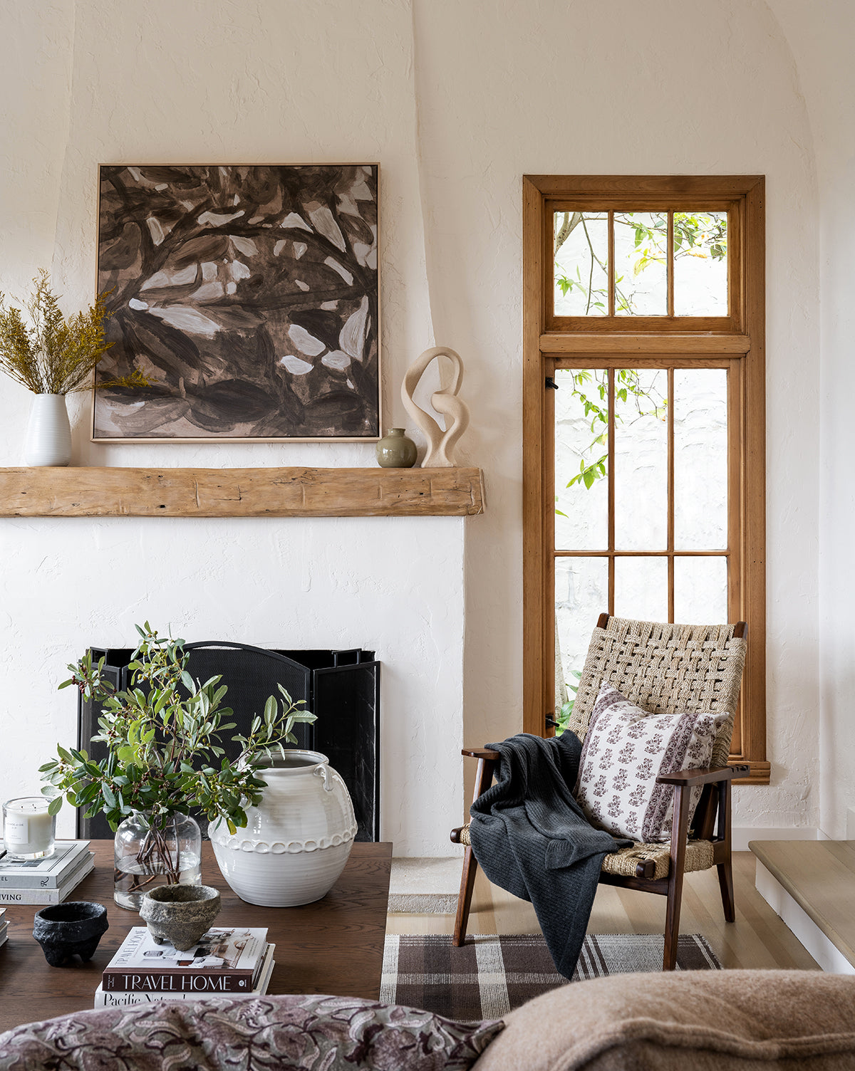 A cozy living room with neutral decor features a wooden window, a woven chair, rustic mantel topped with Light of Summer by Cheryl Clinton, and a wooden coffee table decorated with books, greenery, and ceramics.