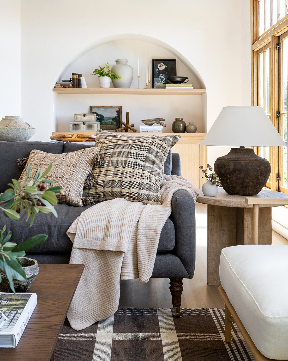 A cozy living room features a gray sofa with plaid and beige pillows, a cream throw, wooden shelves with books and plants, and the Gannon Table Lamp with a fabric shade on a side table near large sunlit windows by McGee & Co.
