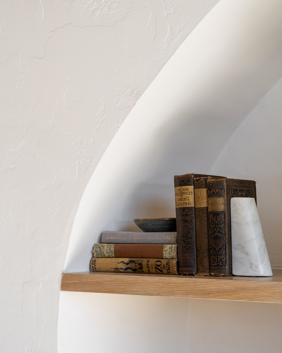 A wooden shelf set in a white textured wall holds vintage books, a small bowl, and the Tapered Marble Bookends (Set of 2). The minimalist scene is softly lit.