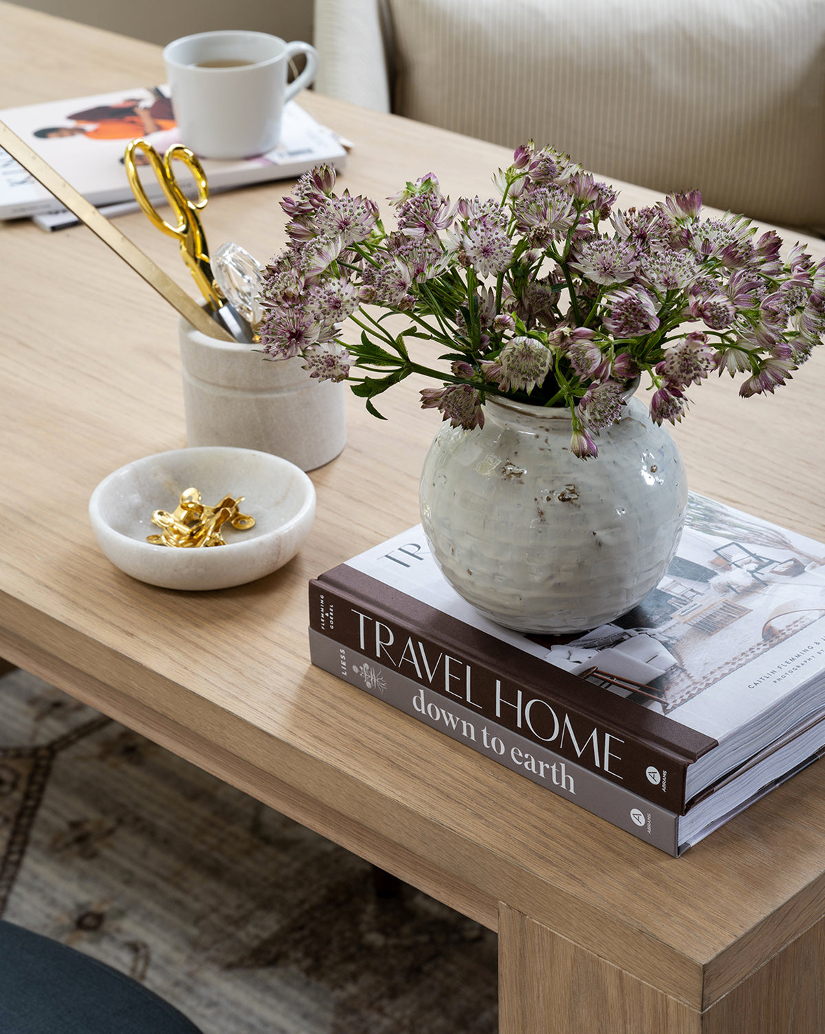 A Barton Sandstone Canister sits on a wooden coffee table beside a vase of purple flowers, two stacked books, a marble bowl with gold clips, gold scissors, and coffee. A sofa with beige cushions is in the background.