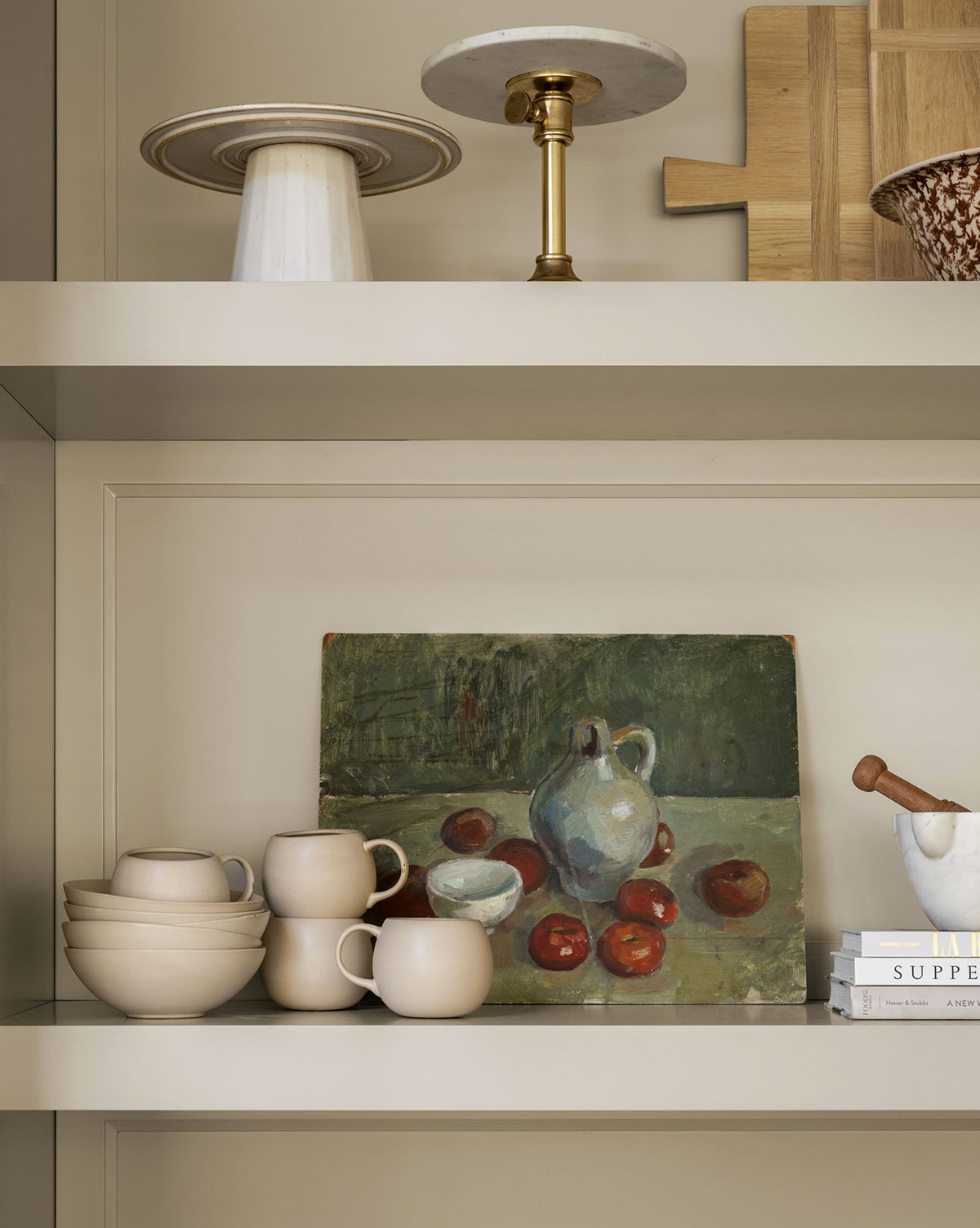 Neutral shelves display glazed stoneware, including the Luana Bowl, cups, a marble cake stand, cutting boards, books, a mortar and pestle, and a still-life painting—creating a modern space with neutral charm, by McGee & Co.