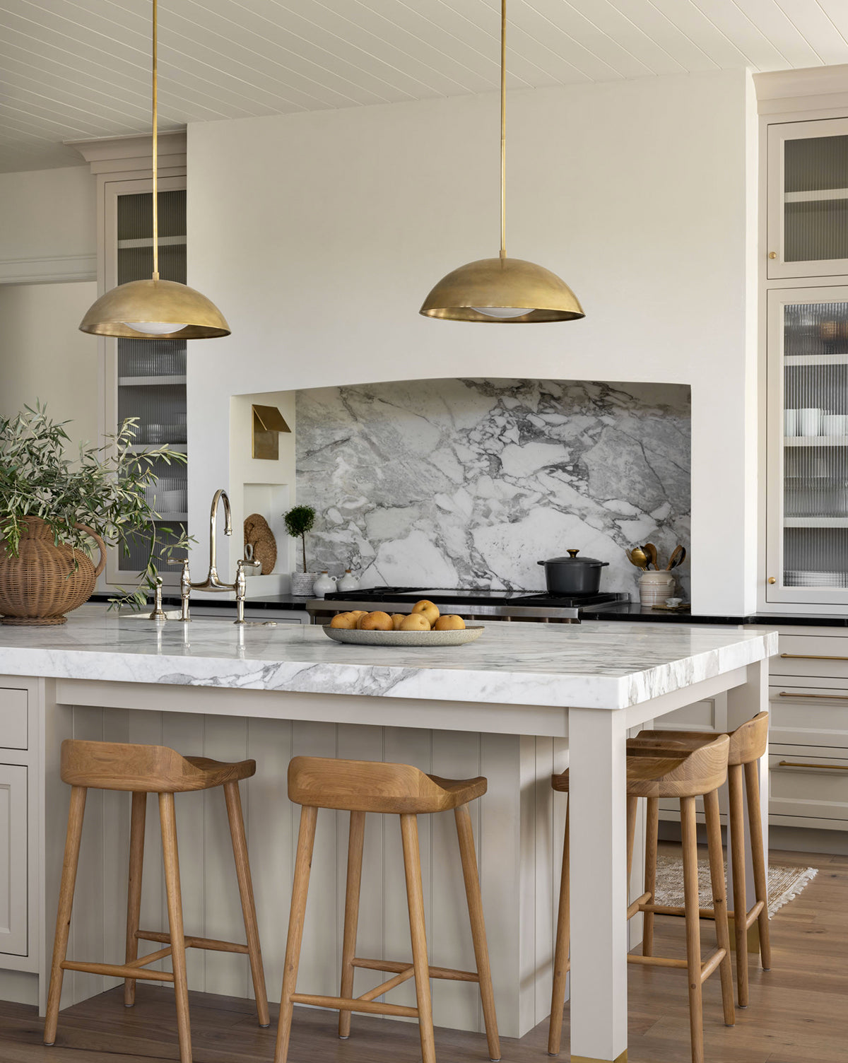 Modern kitchen with a marble island, Makerspalm Portland Counter Stools, gold pendant lights, and a marble backsplash. A bowl of lemons sits on the counter, while a potted plant accents the islands corner. McGee & Co.
